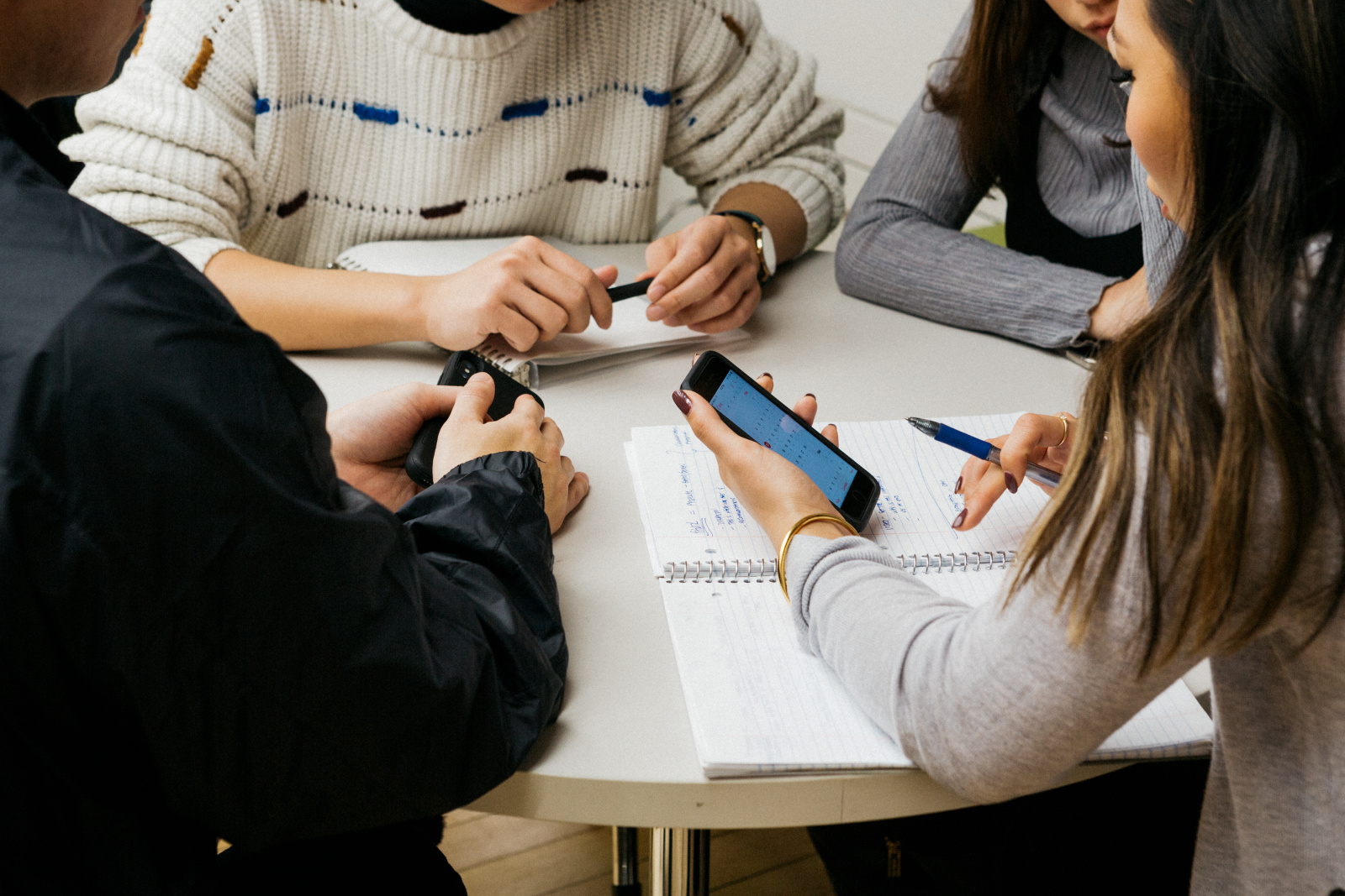 A group of 4 students sit around a table discussing something one of them is showing on their phone. There is an open notebook on the table with notes.