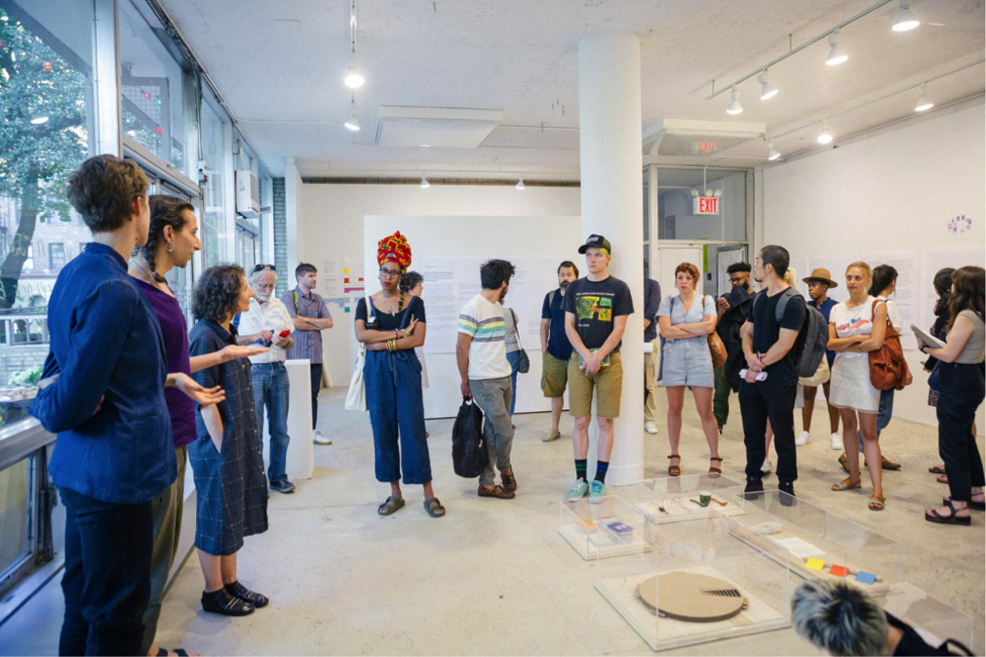 A group of a few dozen people of different ages and demographics stand in a gallery space around plexiglass containers on the floor containing objects. Their attention is on a person speaking.