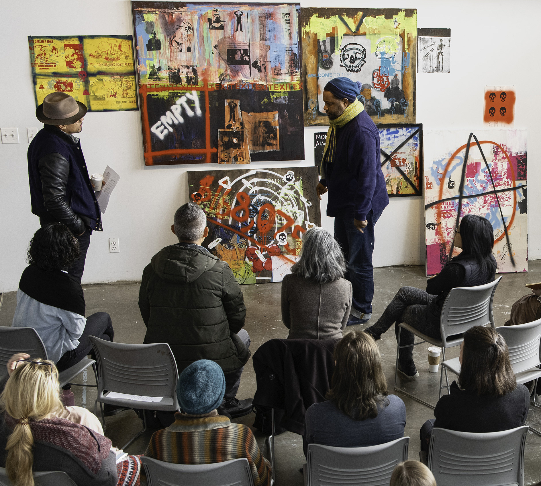 A critique of artwork, happening in a white walled room, with rows of chairs containing students and staff