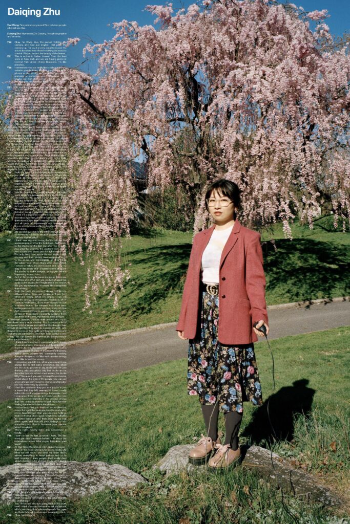 A girl stand in a field of grass with a weeping willow behind her. She is wearing a red cardigan and black pants.