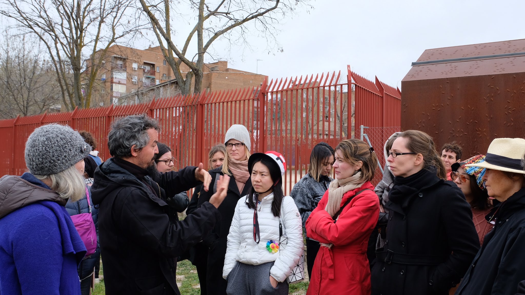 A group of students outside are listening to an instructor speak. Behind them is a red metal fence and some trees.