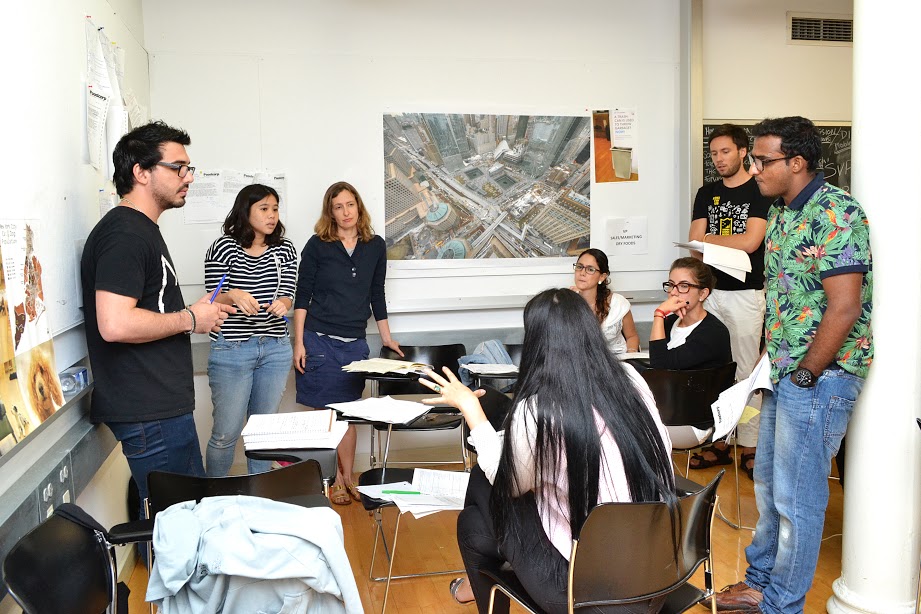 A group of students in a classroom, some sitting, some standing. One is speaking and gesturing with their hands. The other students are listening to them. There are diagrams and schematics on the walls.