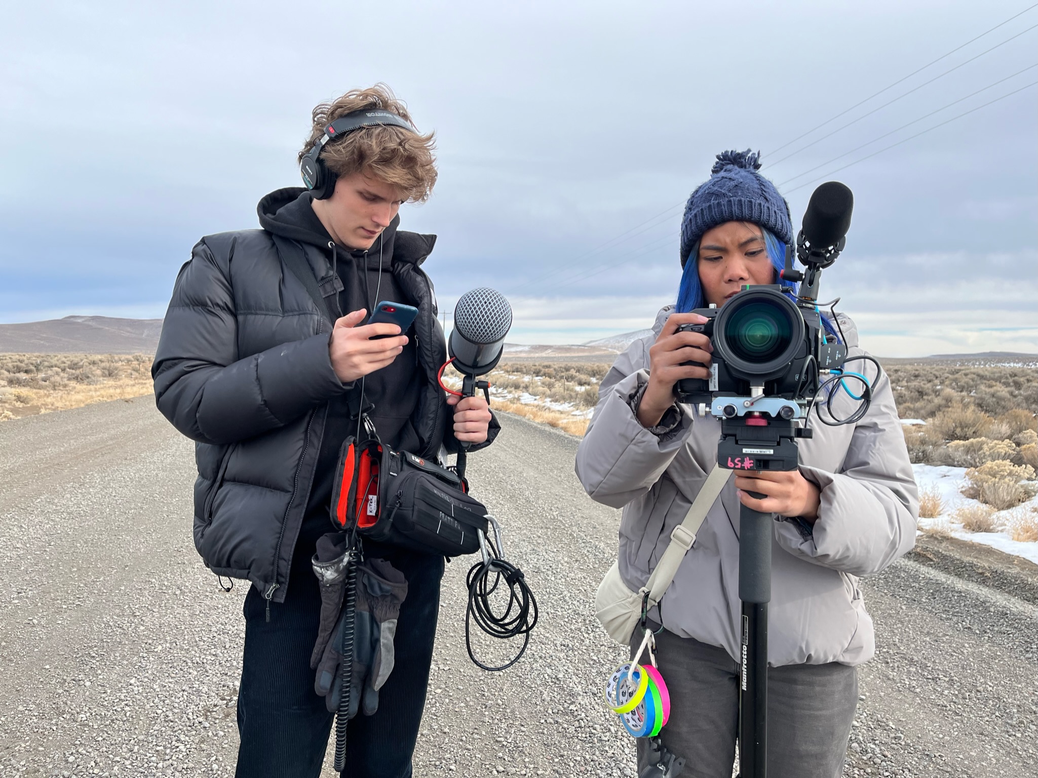 Two students stand on a road with a snowy field expanse around them. They are handling video and sound capturing equipment.