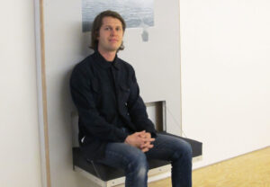 image of Grayson Cox, Adjunct Associate Professor, seated in a chair with industrial, postmodern construction