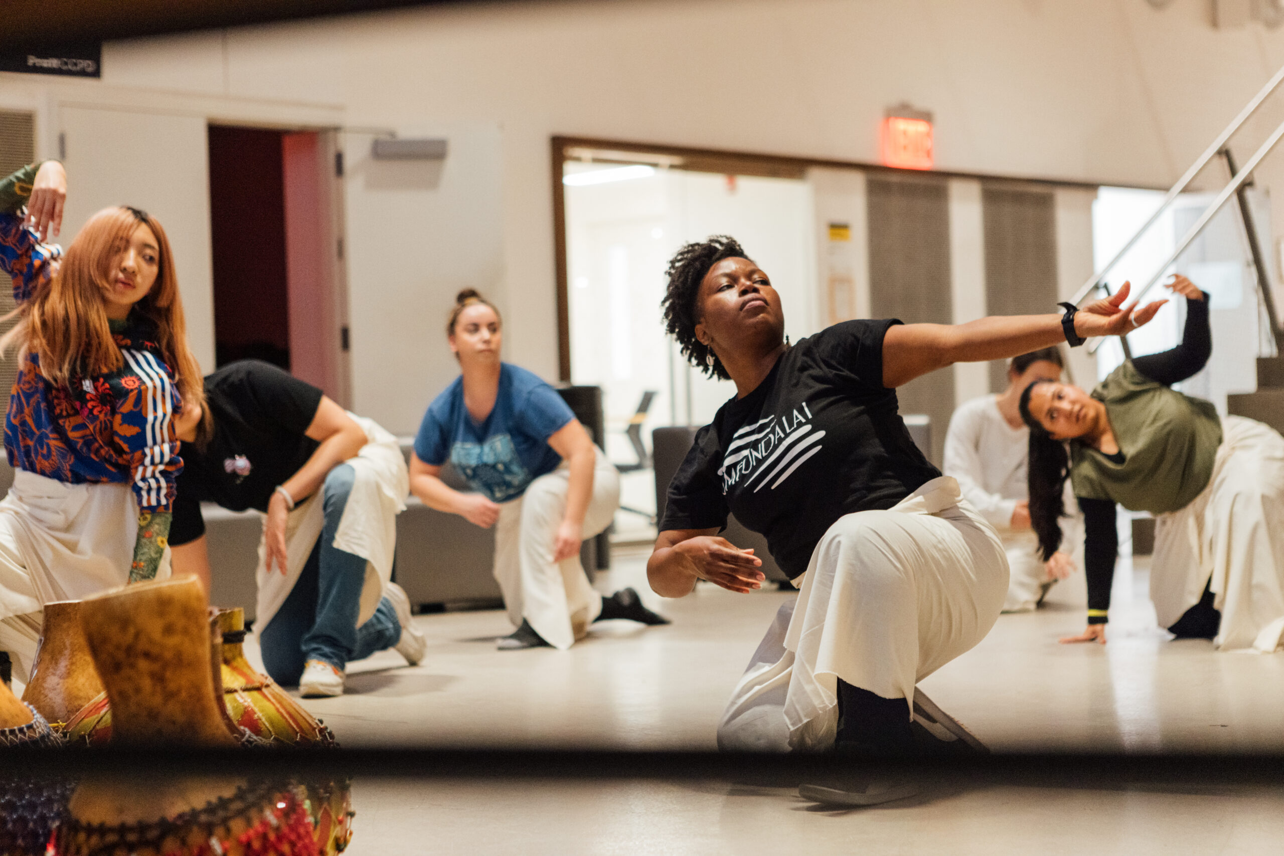 A group of people participate in a dance class, performing a low, dynamic dance move with one arm extended outward. The dancers, including both men and women, wear t-shirts and white wraps around their waists. They are in a modern, spacious room with bright lighting and visible background elements such as doors and stairs. The mood is focused and intense, with each dancer engaged in their movement. The foreground includes colorful gourds placed on the floor, adding to the vibrant atmosphere.