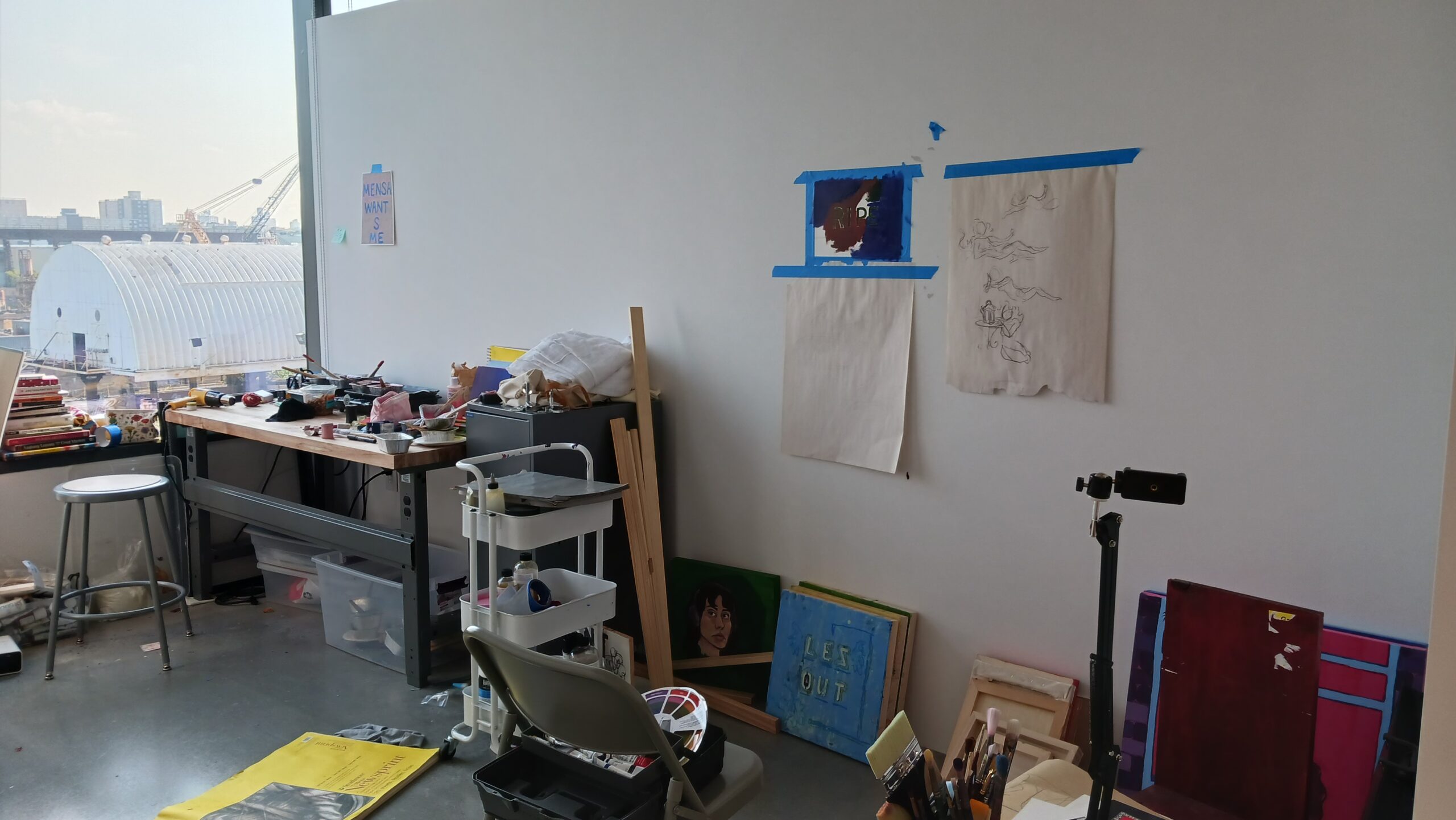 An artist's studio with a window view of an industrial area. The space contains a wooden workbench cluttered with art supplies, small tools, and unfinished pieces. Paintings and sketches are taped to the wall.