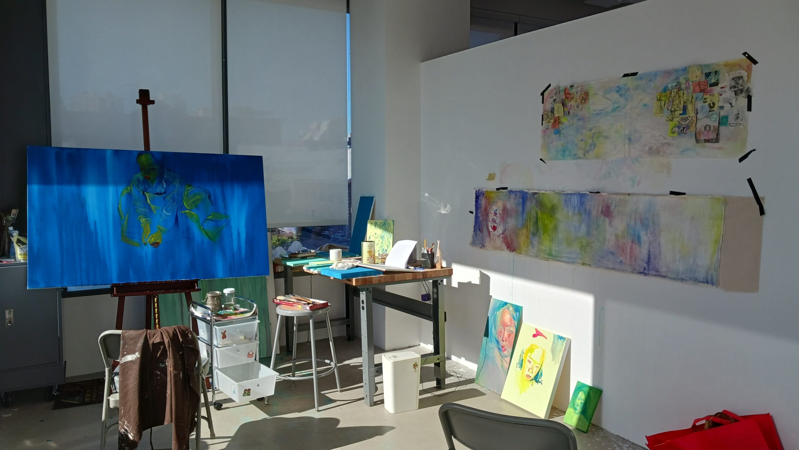 Another view of the artist's studio with an emphasis on a large canvas painting on an easel, surrounded by other artworks and tools. The room is well-lit, featuring expansive windows and a clean, organized workspace.