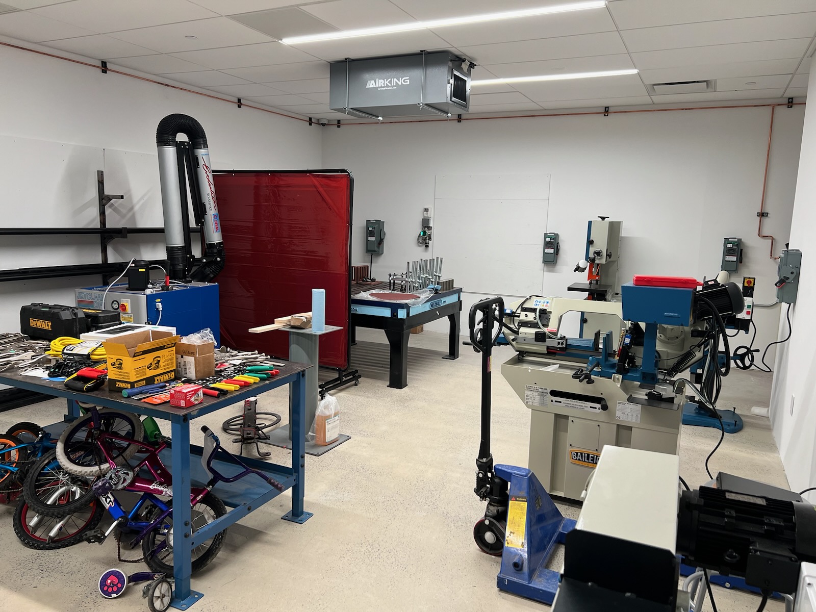 A workshop equipped with various tools and machinery, including a central workbench with clamps and hand tools, a red welding screen, and a collection of bicycles stored underneath a work table. Industrial machines are visible in the background.