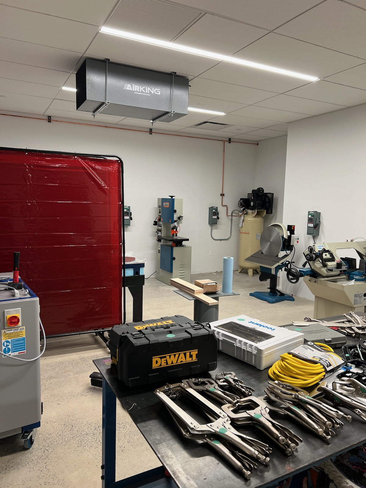 A woodworking and metalworking shop featuring multiple machines, tools, and equipment. The image includes workbenches with vices, a red welding screen, a metal lathe, and other machinery organized across the room. A black air filtration unit is mounted to the ceiling.