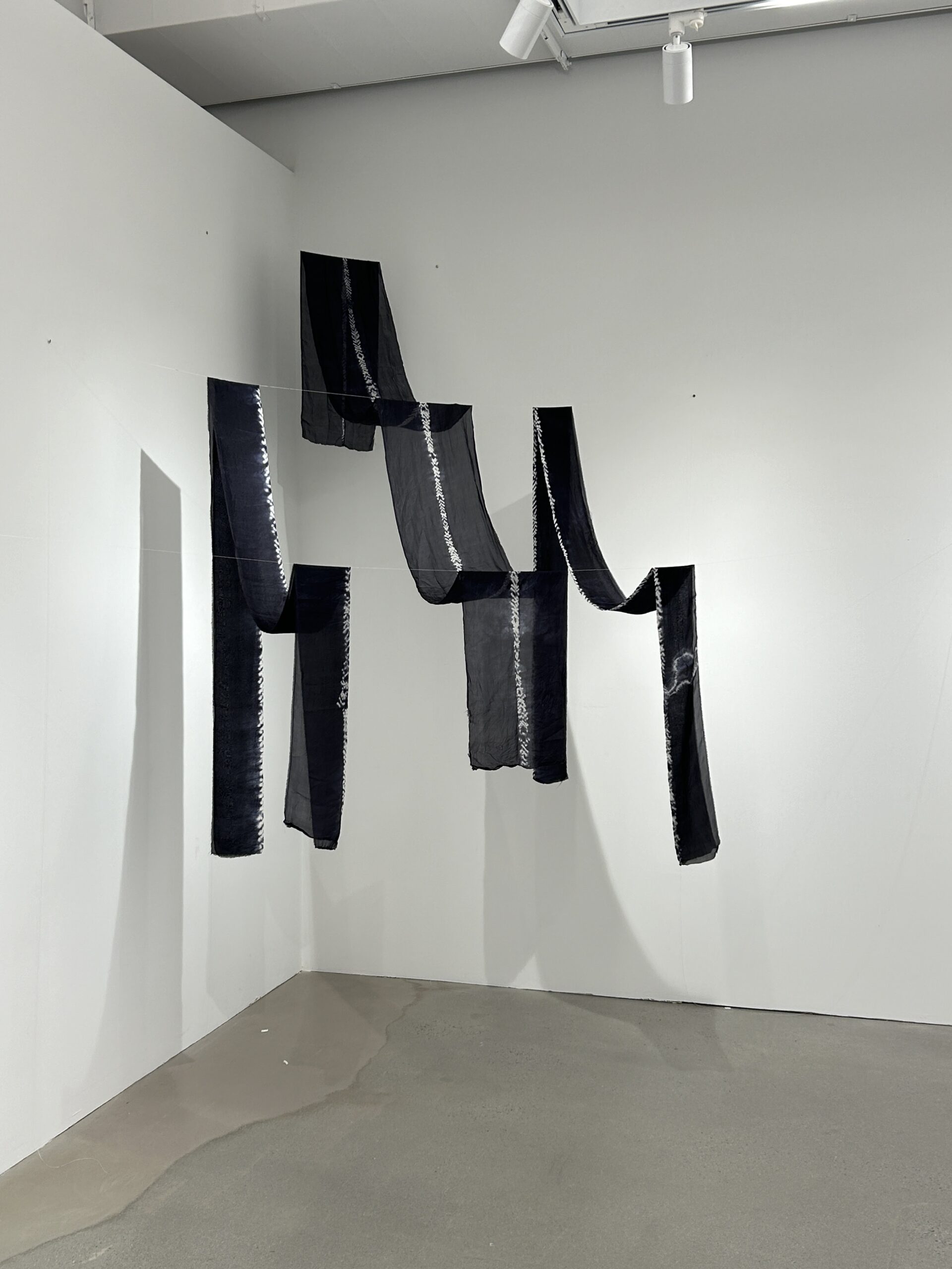A contemporary art installation featuring dark, semi-transparent fabric pieces suspended in the air. The arrangement creates a flowing, layered appearance against the white gallery walls.