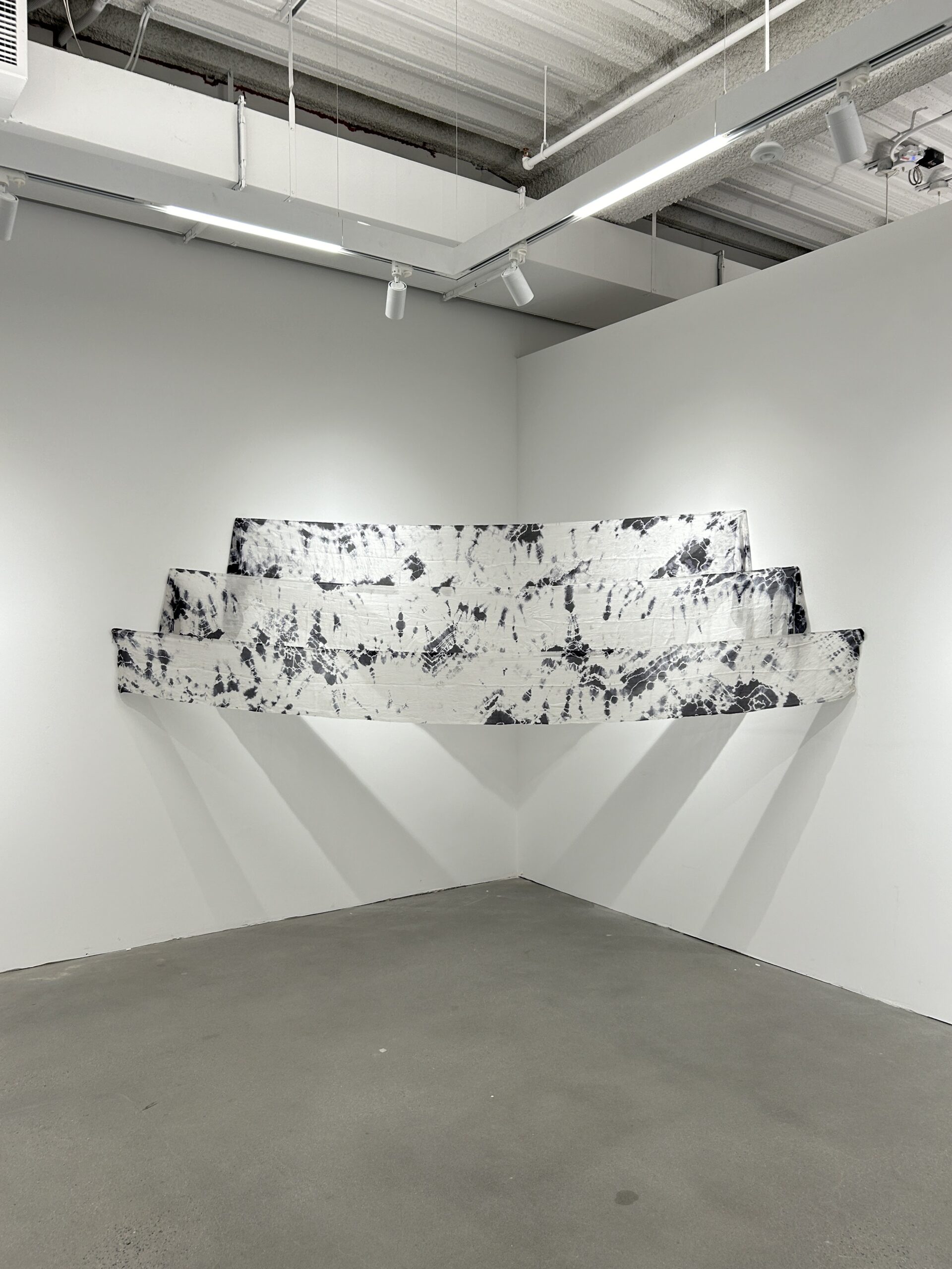 A minimalist art installation in a gallery setting, showcasing a layered white fabric structure with black tie-dye patterns. The installation is mounted in a corner with soft lighting emphasizing the textures.