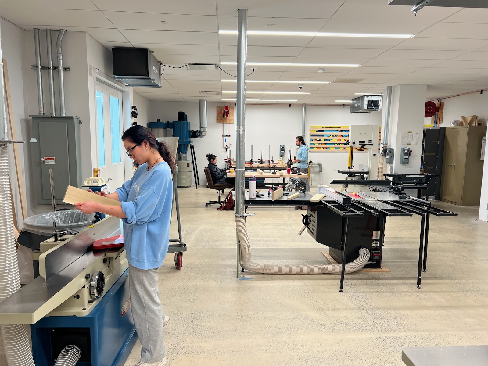A woodworking workshop with multiple tables, machinery, and tools. A person is sanding a wooden piece on a jointer while others work at various stations. The room has a clean and organized layout with large windows allowing natural light.