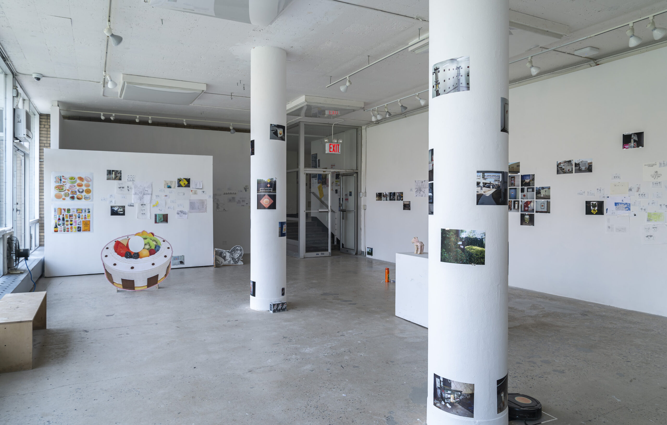 An open gallery space with a variety of photos, sketches, and mixed media artworks displayed on walls and columns. The room has a casual, exploratory atmosphere with natural light coming through large windows.