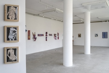 An art gallery installation focused on equine themes, featuring a collection of horse sketches, paintings, and photographs. A pedestal in the center supports an open book with additional horse illustrations. The artworks are arranged in an organic, clustered manner, with some pieces extending onto the floor. A large paper bag labeled 