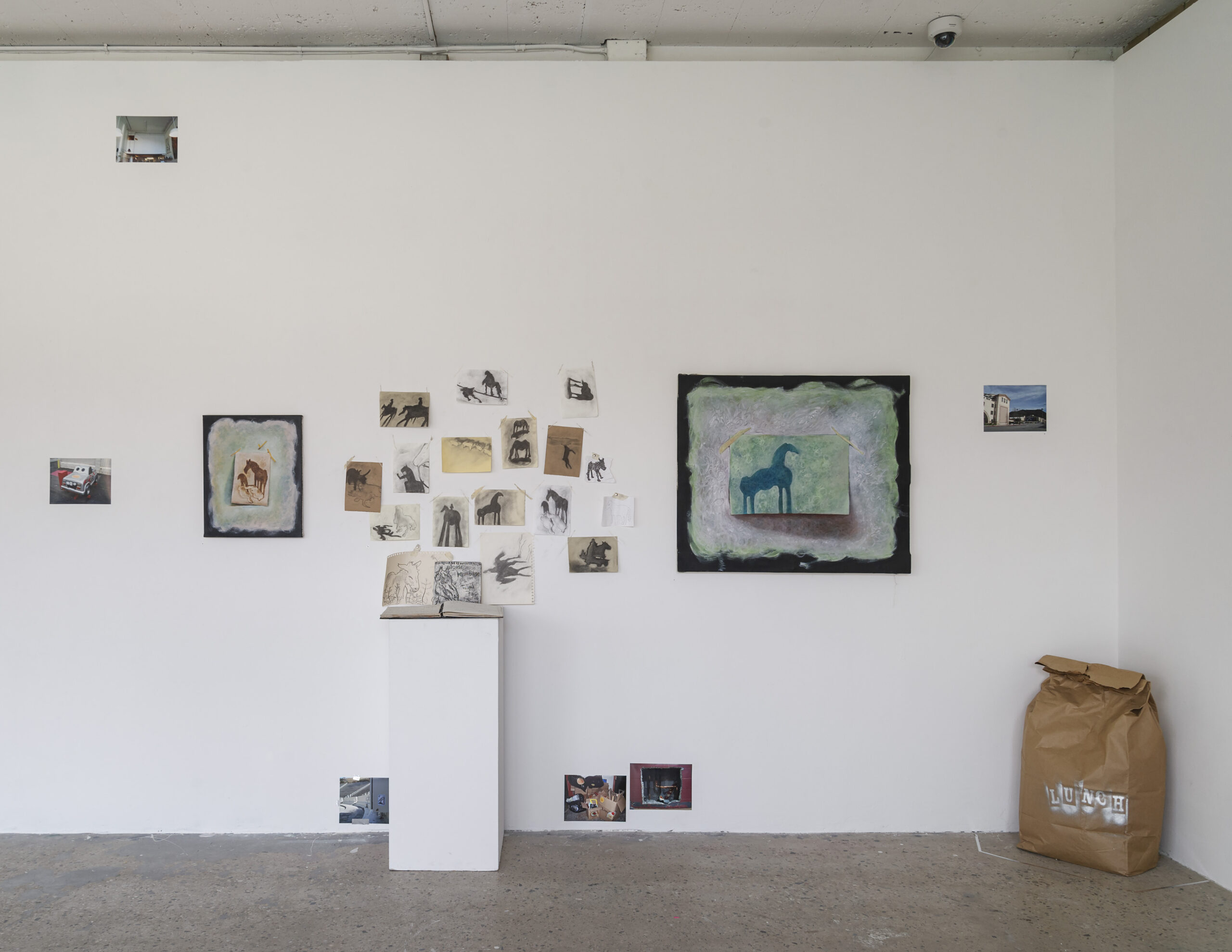 A contemporary gallery corner with a series of paintings inspired by urban landscapes and city life. The leftmost painting depicts a subway scene with two figures sitting in a dimly lit train. Other pieces include abstract neon-colored compositions, architectural depictions, and nighttime cityscapes. The exhibition space is well-lit, with a polished concrete floor and a window allowing natural light to enter.