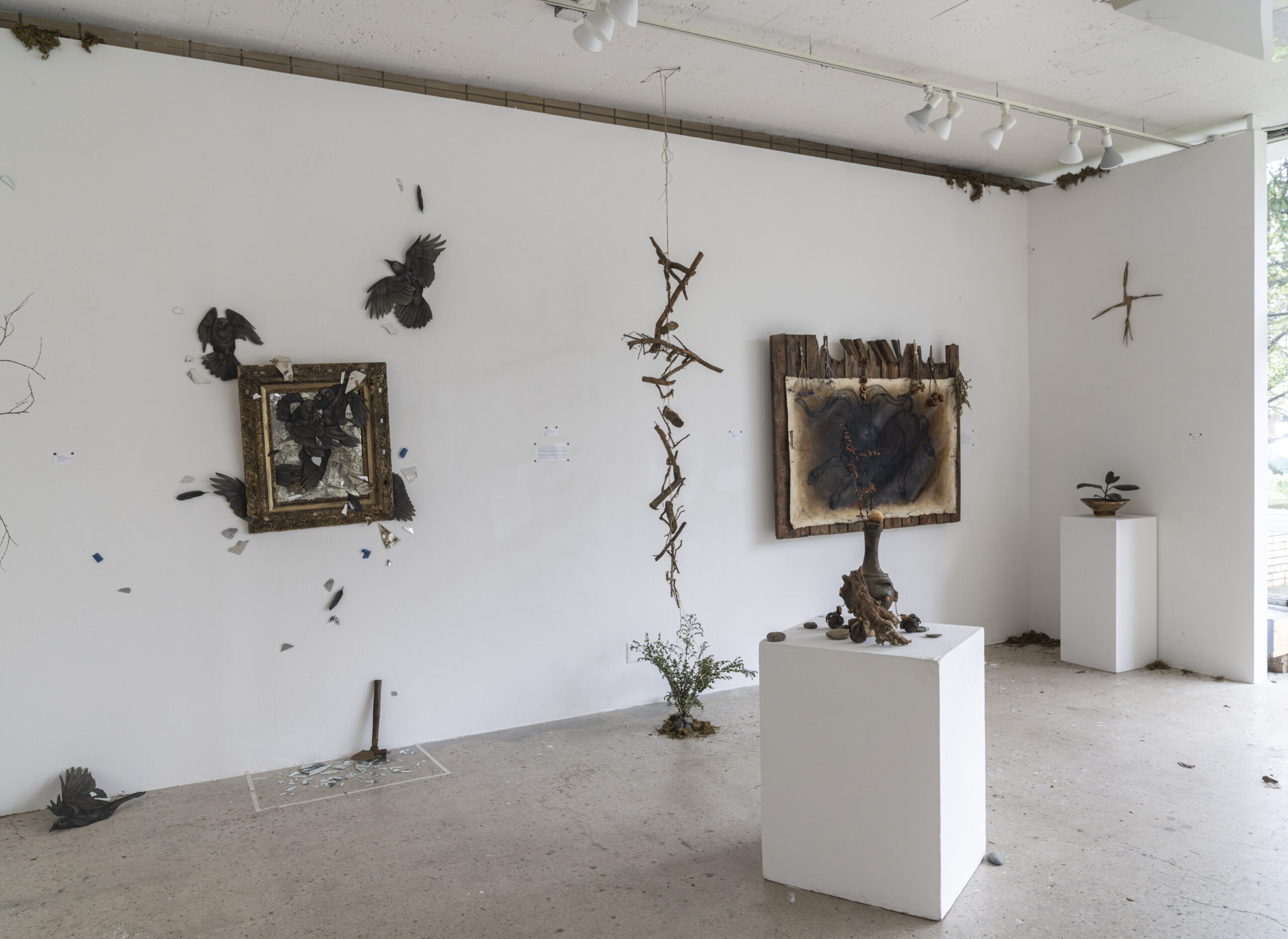 A spacious gallery exhibition featuring a series of small and medium-sized artworks mounted along a long white wall. The room is divided by white pillars, and the artworks range from black-and-white portrait sketches in wooden frames to colorful figurative and abstract compositions. The polished concrete floor and high ceilings contribute to the minimalist aesthetic.