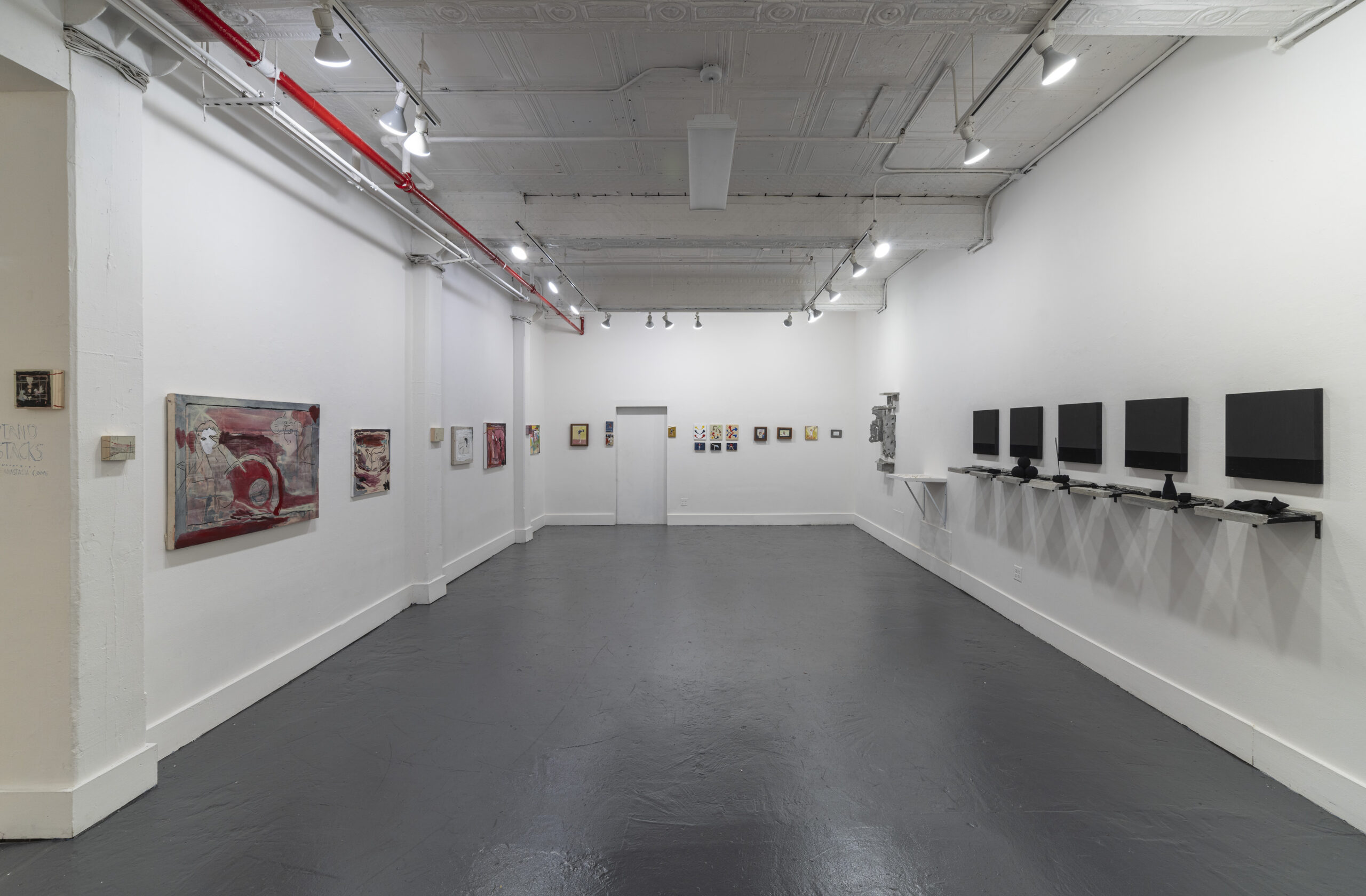 A contemporary art gallery room with a wooden floor and white walls, displaying several small paintings arranged in a linear fashion. The paintings vary in subject matter and style, with some featuring abstract elements and others depicting more figurative scenes. The room is illuminated by multiple overhead spotlights.