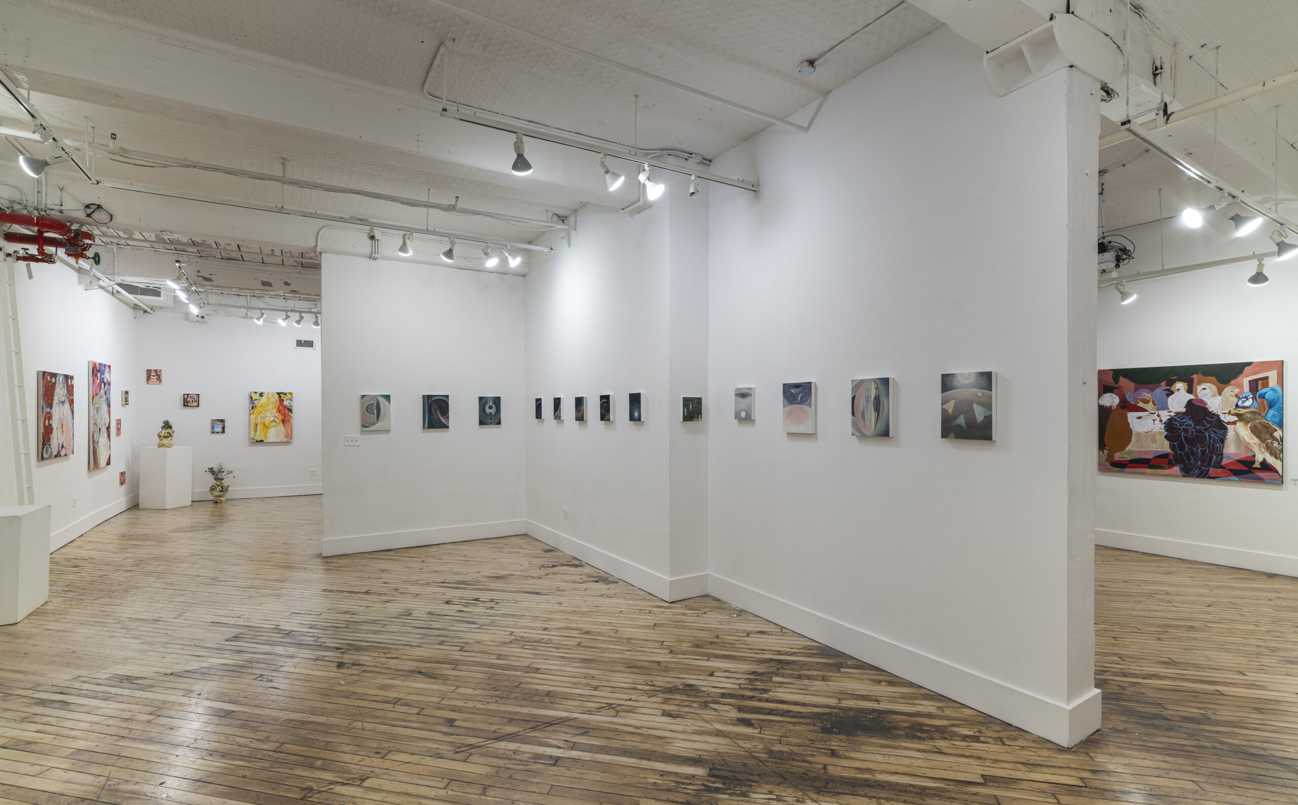 A contemporary gallery space featuring a diverse range of artwork. The left side showcases a series of brightly colored geometric paintings with layered, puzzle-like patterns, while the right side displays black-and-white surrealist prints. The gallery's wooden floor and white walls provide a neutral backdrop for the vibrant artwork.