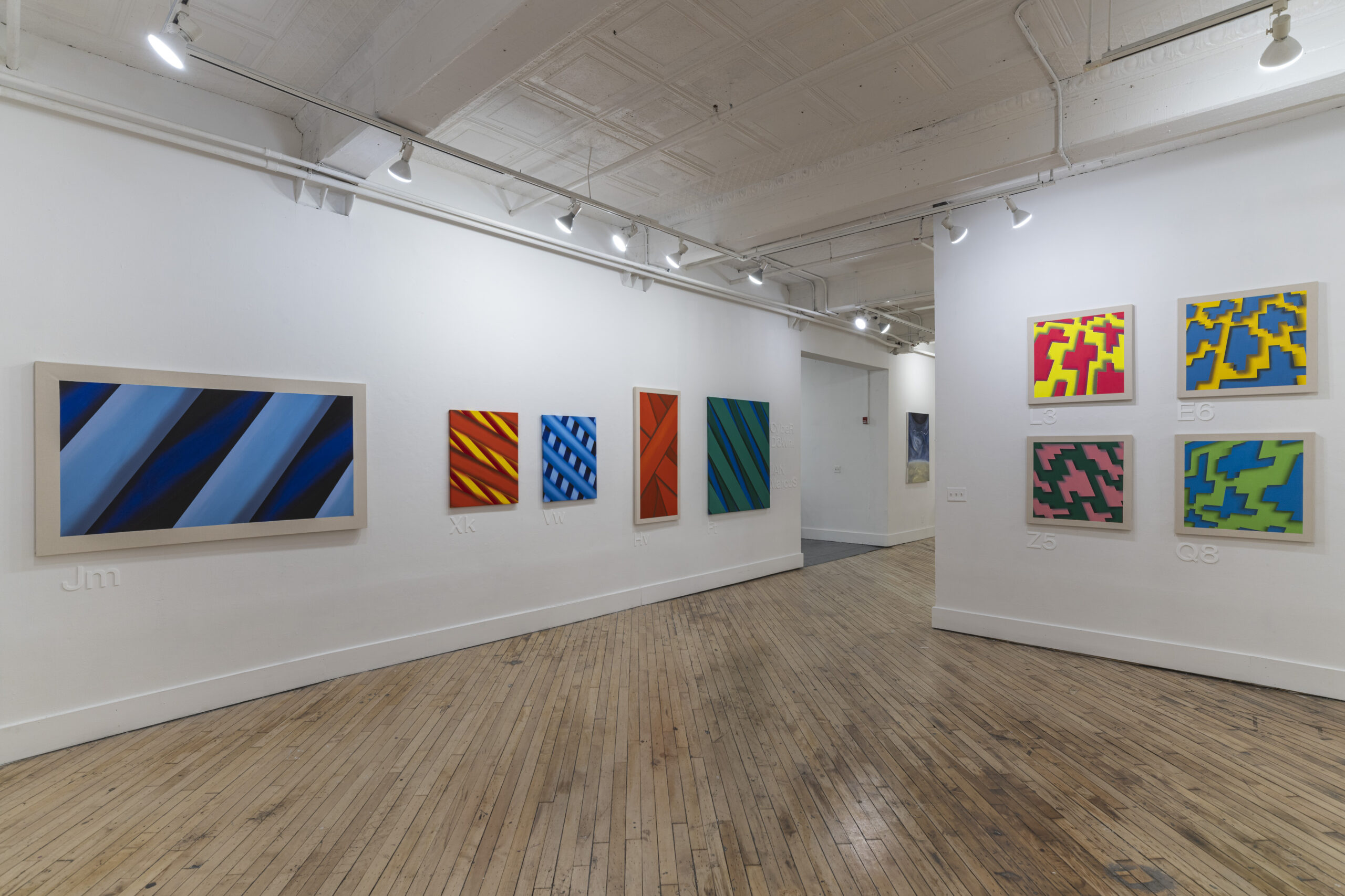 A spacious art gallery with a polished wooden floor and white walls, featuring a variety of paintings in different styles. The foreground presents four colorful geometric paintings, while the background has black-and-white graphic works depicting surreal objects. Bright spotlights highlight the artwork on display.