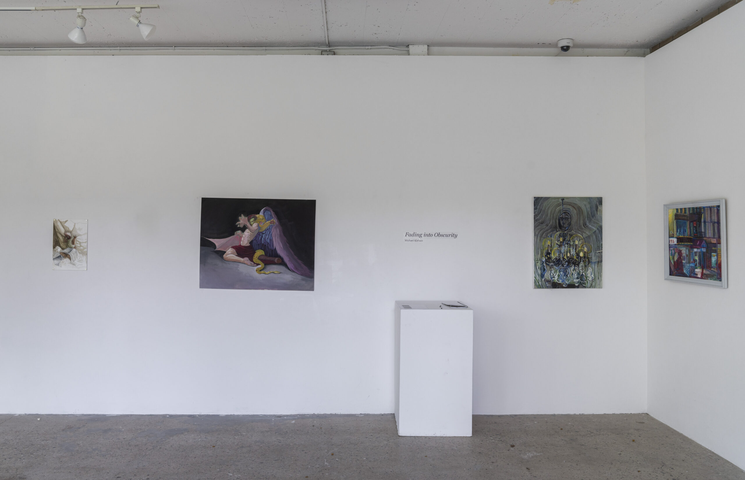 An art gallery with a white wall and a concrete floor, displaying four paintings of varying styles. The centerpiece is a large painting depicting a figure draped in purple fabric interacting with a snake. To the right, two framed paintings feature an abstract portrait with candlelight and a colorful interior scene. A small white pedestal with a notebook is positioned beneath the exhibition title, 