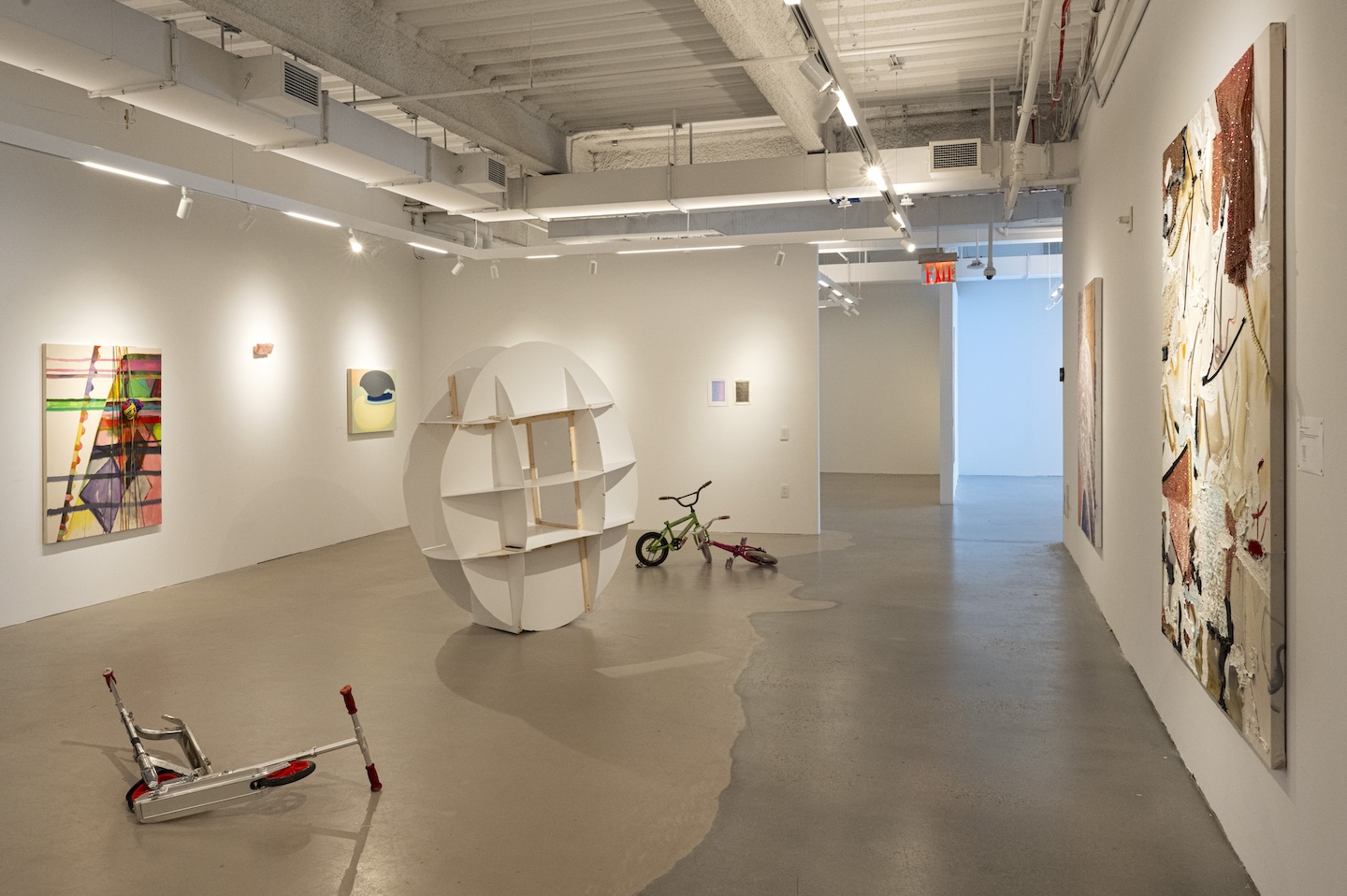A contemporary gallery exhibition featuring abstract paintings with a variety of textures and colors. On the left, a large textile-based artwork with intricate symmetrical patterns is displayed, while on the right, a vibrant diptych with bold yellow, red, and blue brushstrokes is mounted. In the foreground, a sheer, blue-tinted fabric hangs from the ceiling near a large window, allowing natural light to interact with the exhibit.