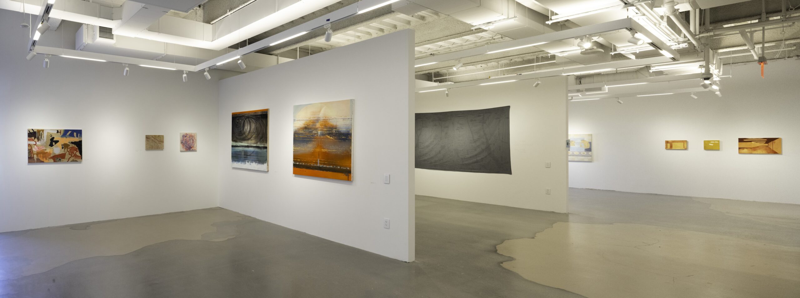 A gallery corner displaying a mix of abstract paintings and conceptual art pieces. Three small paintings with warm, earthy tones and geometric shapes are mounted on the left wall. A tall, white pedestal in the center holds a small glass with a dark liquid. To the right, a black pedestal houses an enclosed religious icon painting inside a glass case, adding a historical contrast to the contemporary setting.