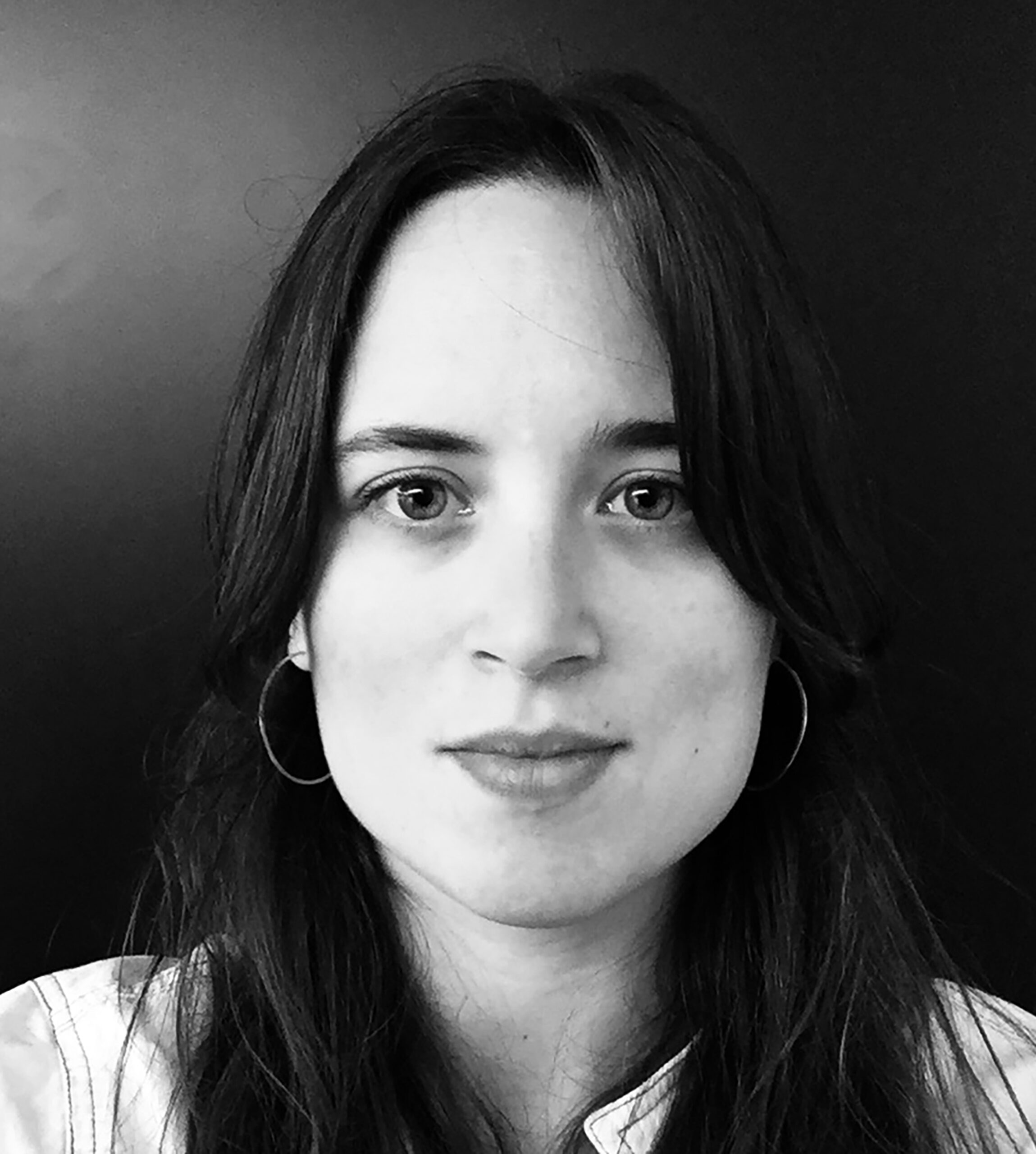 A black and white portrait photograph of a Caucasian woman with dark hair from the shoulders up. Her hair goes past the bottom of the frame. She is centered in the photograph and looks directly at the camera wearing a neutral facial expression.