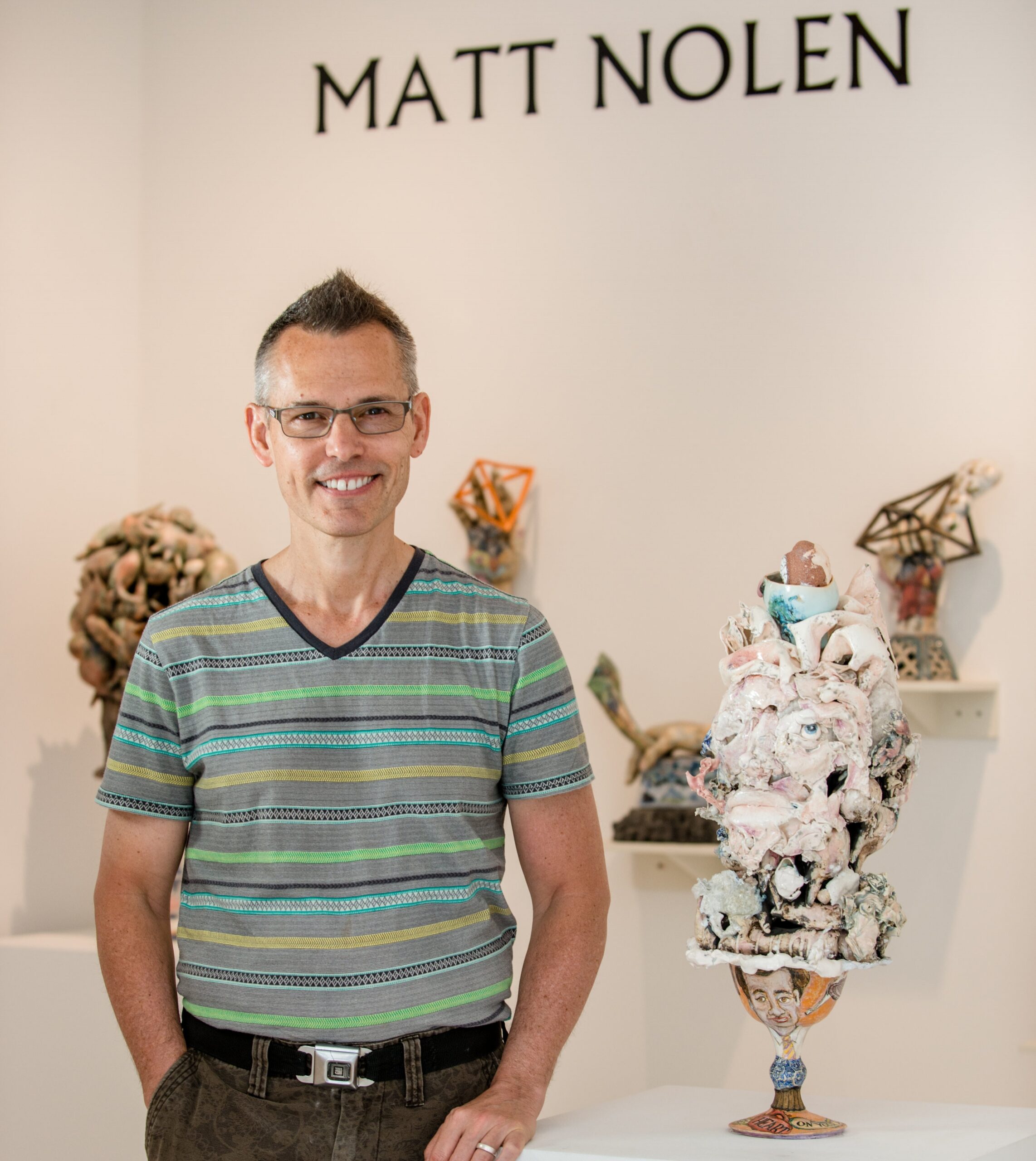 Portrait of Matt Nolen in a white-walled gallery, standing next to a sculpture. His name is written on the wall behind him.
