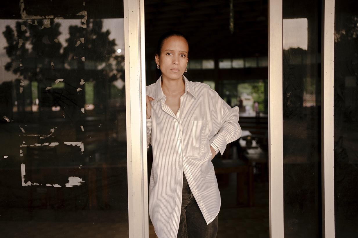 A person stands partially framed by a doorway, looking directly at the camera with a calm, serious expression. They are wearing a long, oversized, light-colored striped button-down shirt and dark pants. One hand is placed on the doorframe while the other rests on their hip. The background shows a dimly lit interior space with large windows, wooden furniture, and outdoor greenery visible through the glass. The lighting is soft and natural, adding a contemplative tone to the image.