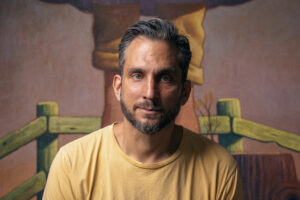 Portrait of Nat Meade in front of part of a painting. He is centered in the frame, looking straight ahead, and wearing a yellow shirt.