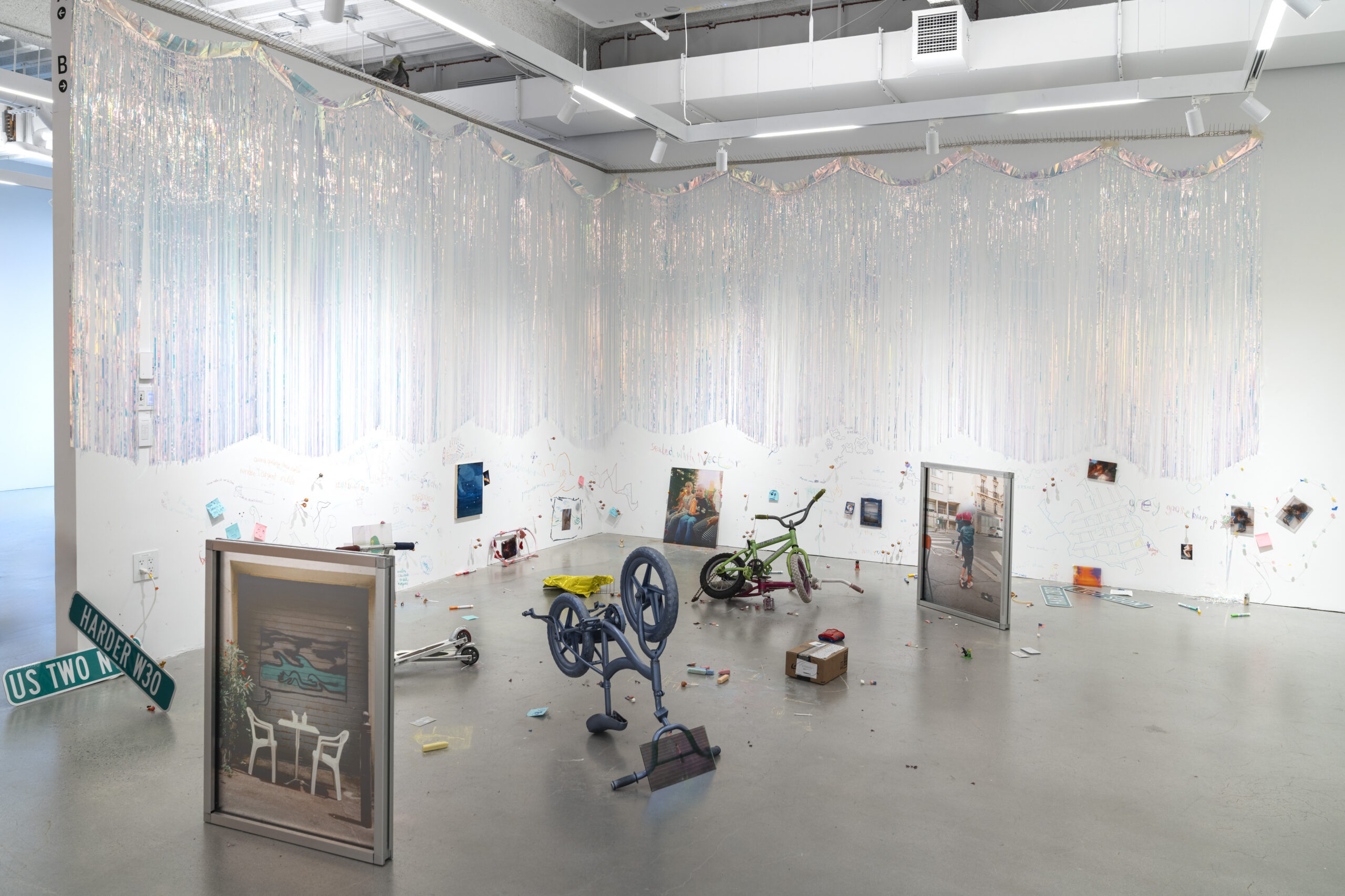 Artwork installation featuring holographic fringe hanging from top of the wall all the way around the gallery booth. On the floor in various locations are vertical framed photographs, a bike, and other objects.