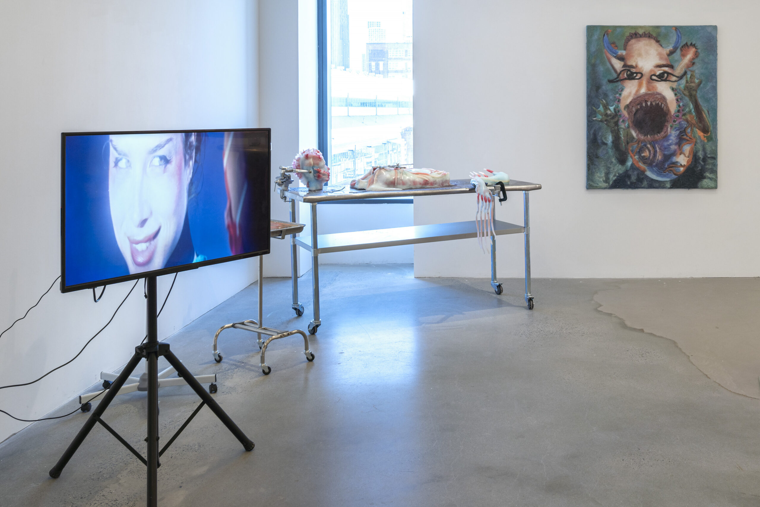 Gallery installation view from left to right: a monitor on a tripod stand playing a film; a metal operating table on which what appears to be a figurative sculpture lays, and a face with an open mouth hanging on the back wall.