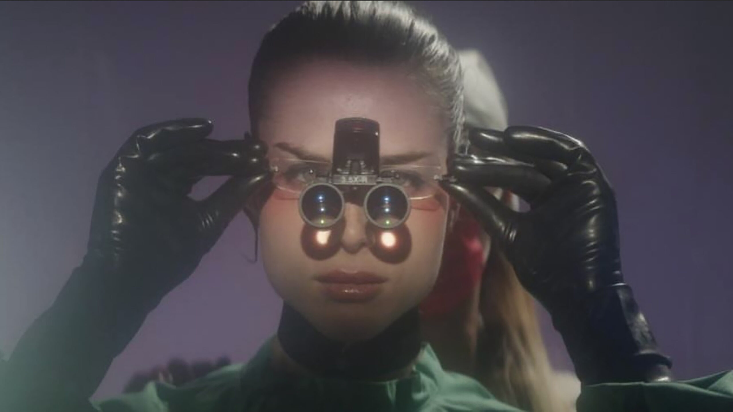 Film still: a person with long hair tied up in a ponytail centered in frame holds a pair of binoculars to their face wearing black gloves