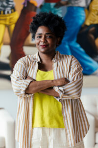 Photo of Sam Vernon with her arms crossed in front of a colorful artwork.