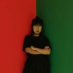 Photo of eri king wearing a black t-shirt and with arms crossed. The background is split horizontally down the center, half red on the left and the other half green on the right.