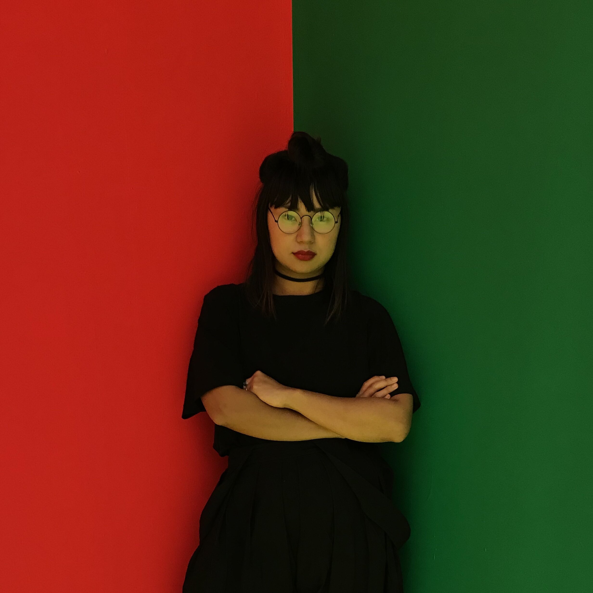 Photo of eri king wearing a black t-shirt and with arms crossed. The background is split horizontally down the center, half red on the left and the other half green on the right.