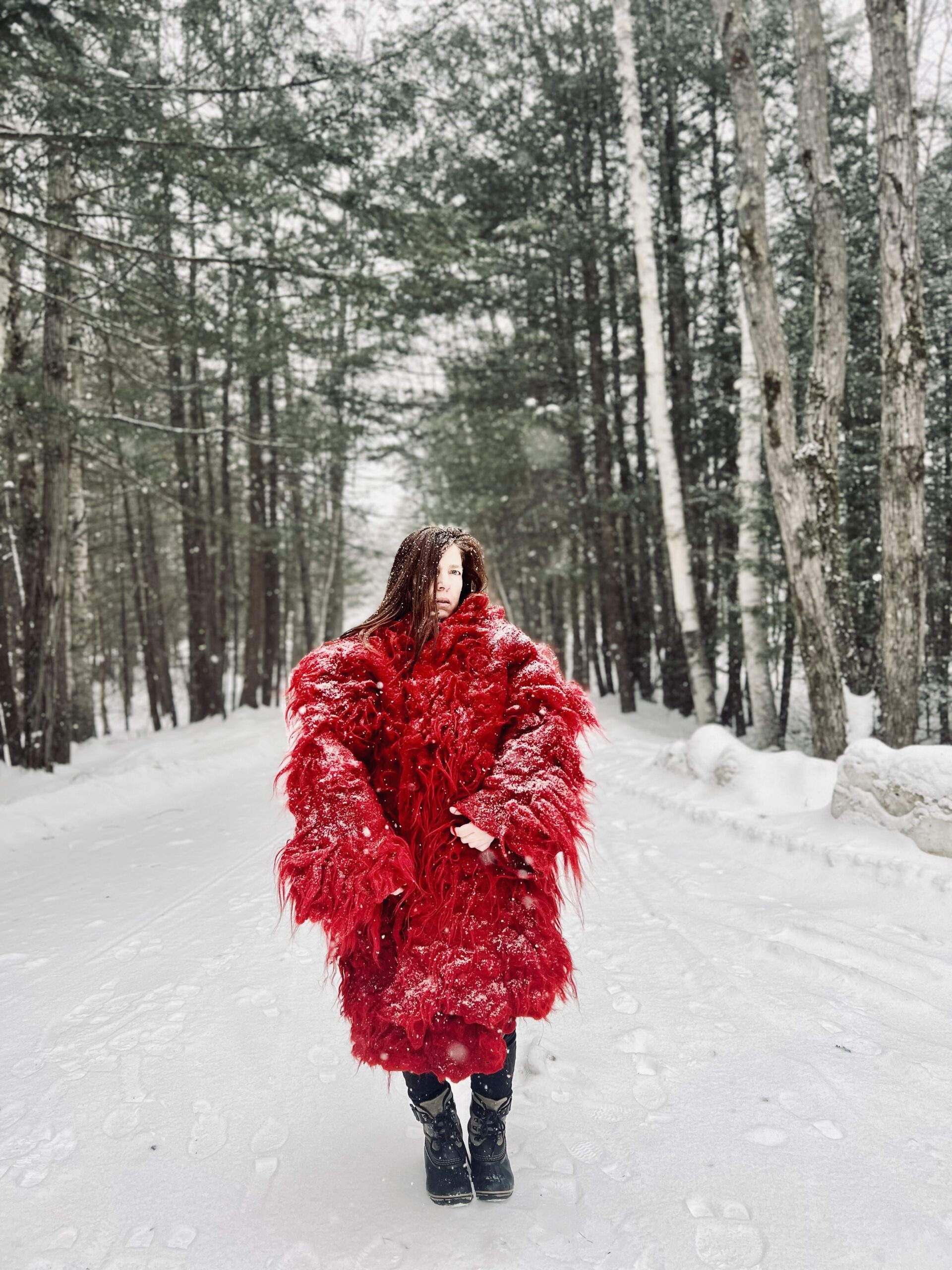 Film still from Hunia-Permission to Be by Brigitta Varadi. The artist stands at the center of a snow-covered road flanked on both side by pine trees, wearing a large red coat.