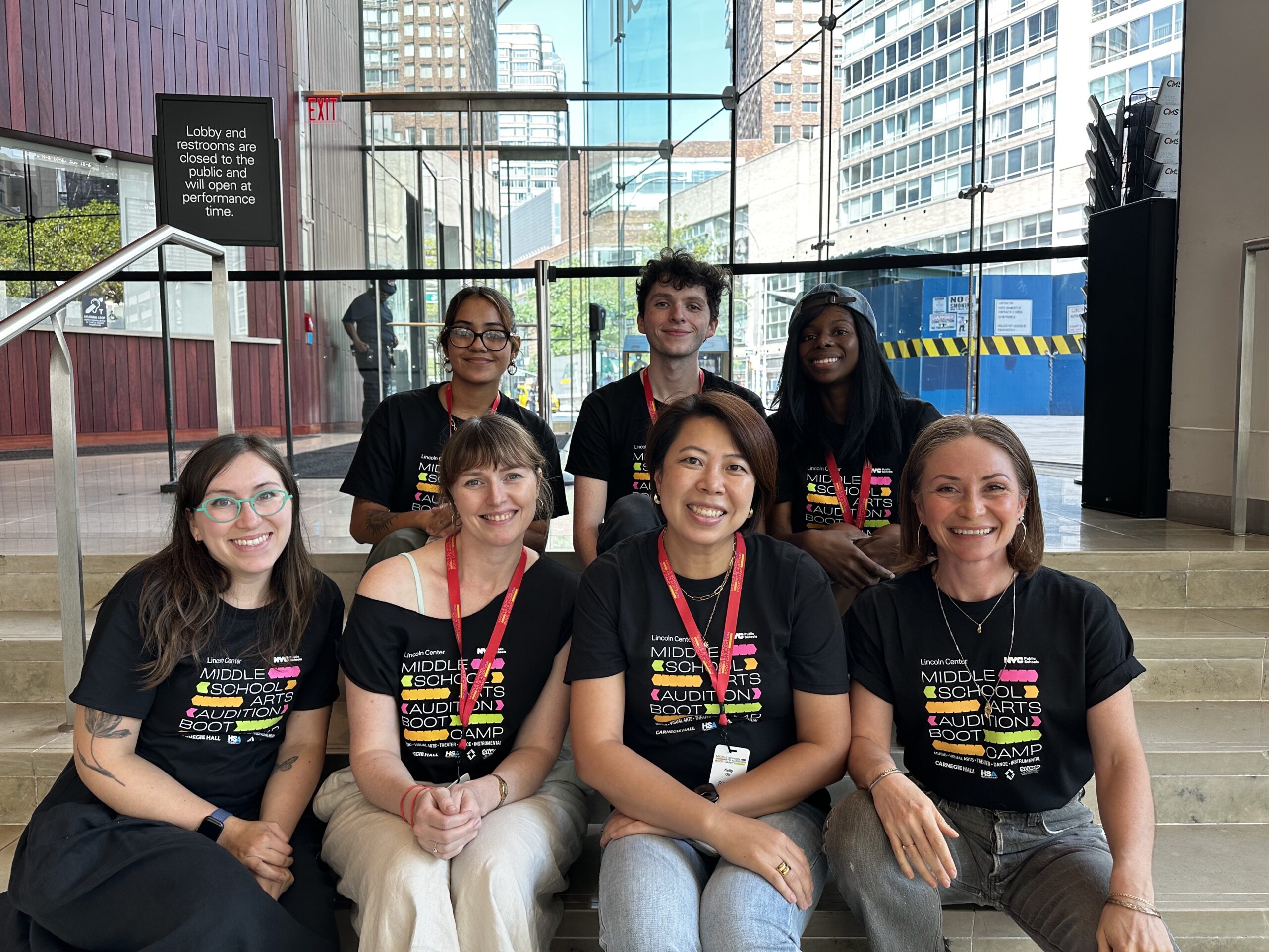 Group photo of six people sitting on indoor steps in a glass-walled atrium. They are smiling and wearing matching black t-shirts with colorful text that reads “Middle School Arts Audition Boot Camp.” A sign in the background indicates that lobby and restrooms open at performance time.