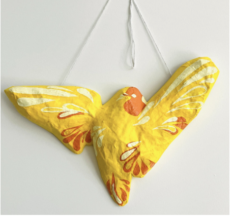 Photograph of a yellow painted paper mache bird sculpture with orange accents, hung by a white string on a wall. Identified as a protection sculpture project example, showing a flying bird shape with wings spread wide.