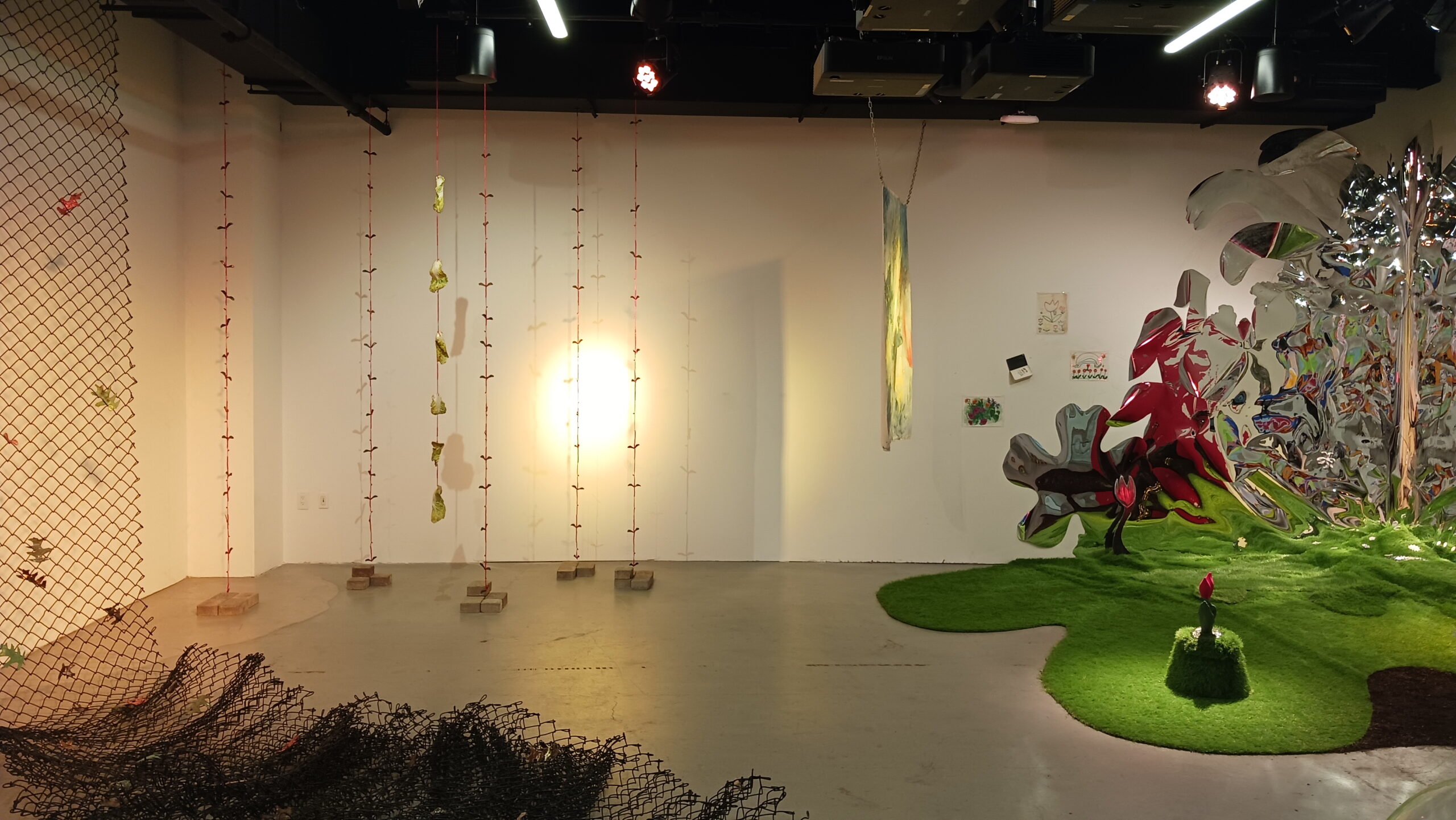 A dimly lit room with a warm yellow light spotlighting on the back wall. To the left of the room is a net with leaves hanging from the ceiling and piling on the floor. In the corner, a series of ropes hang connected to bricks on the floor. A panel also hangs from the ceiling. To the left is a large installation featuring false turf, tulip forms, and rippled silver.