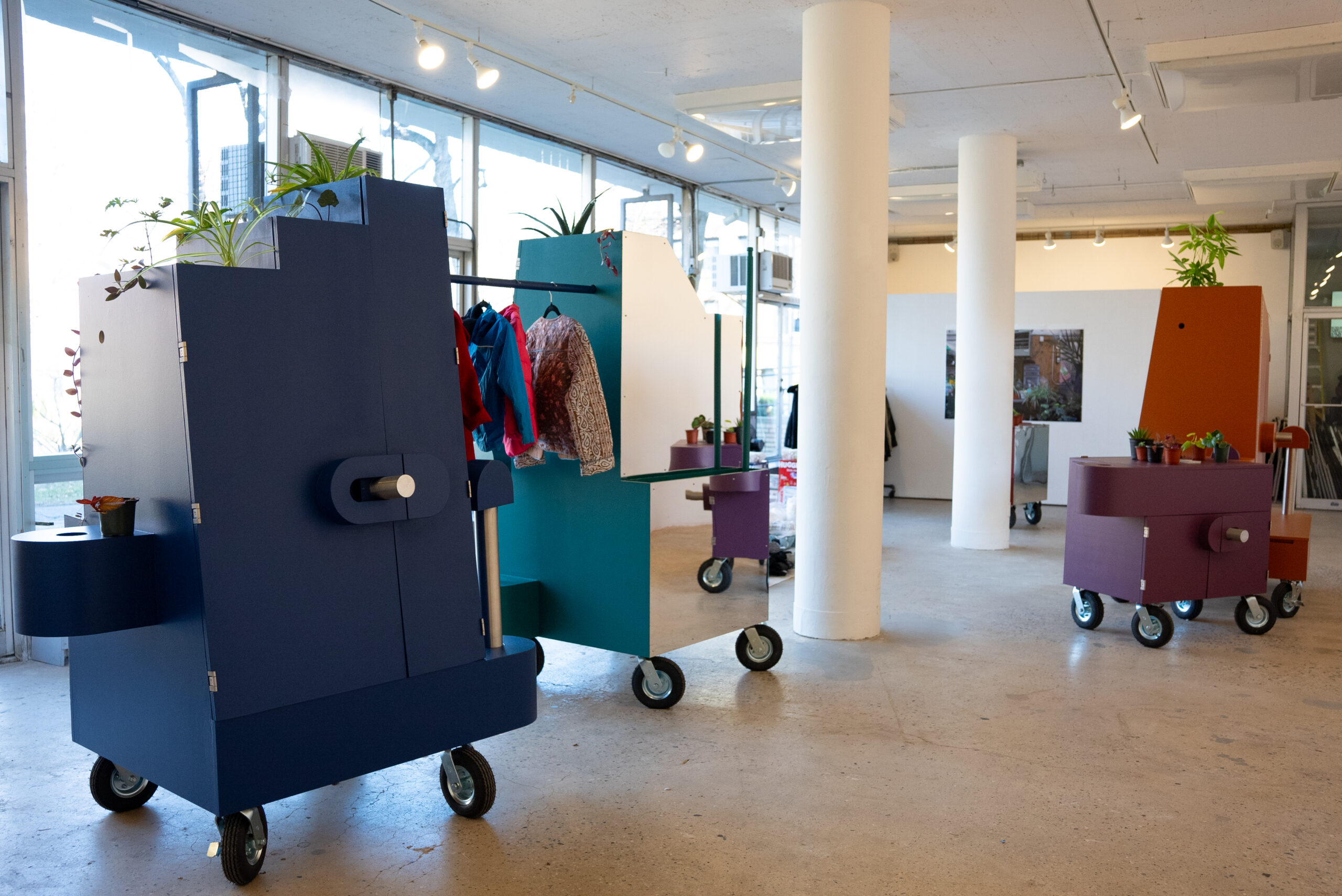 A brightly lit gallery featuring movable, color-blocked, modular sculptures on wheels. One dark blue features a door on the front and is connected with a lighter blue sculpture by a rod, from which clothes on hangers hang. The side of the lighter blue sculpture is reflective. There are plants resting atop these sculptures and elsewhere in the room.