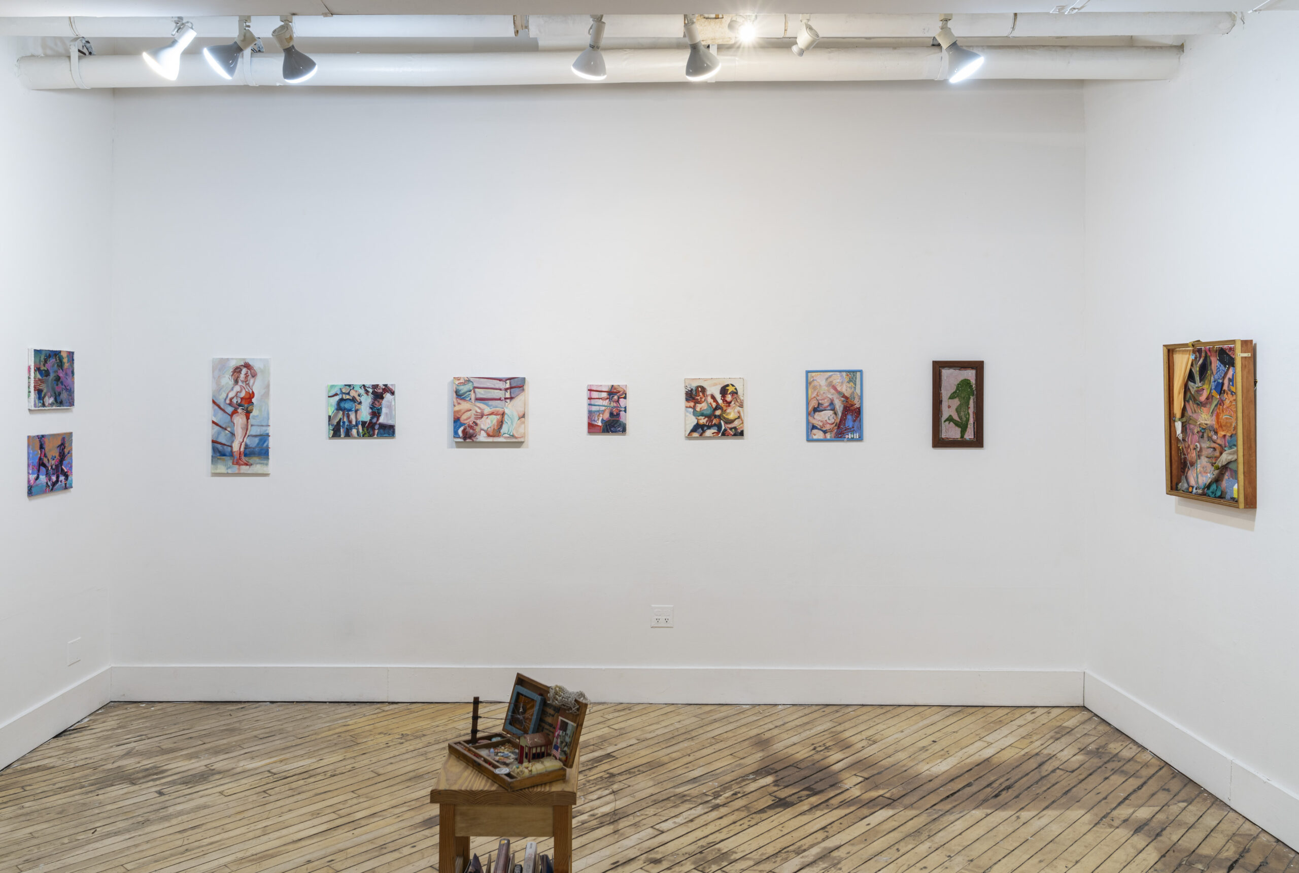 A brightly lit gallery with a row of small paintings across the back wall. Other small paintings hand on the end walls to either side. In the middle of the room toward the foreground of the photo is a sculptural installation featuring miniatures
