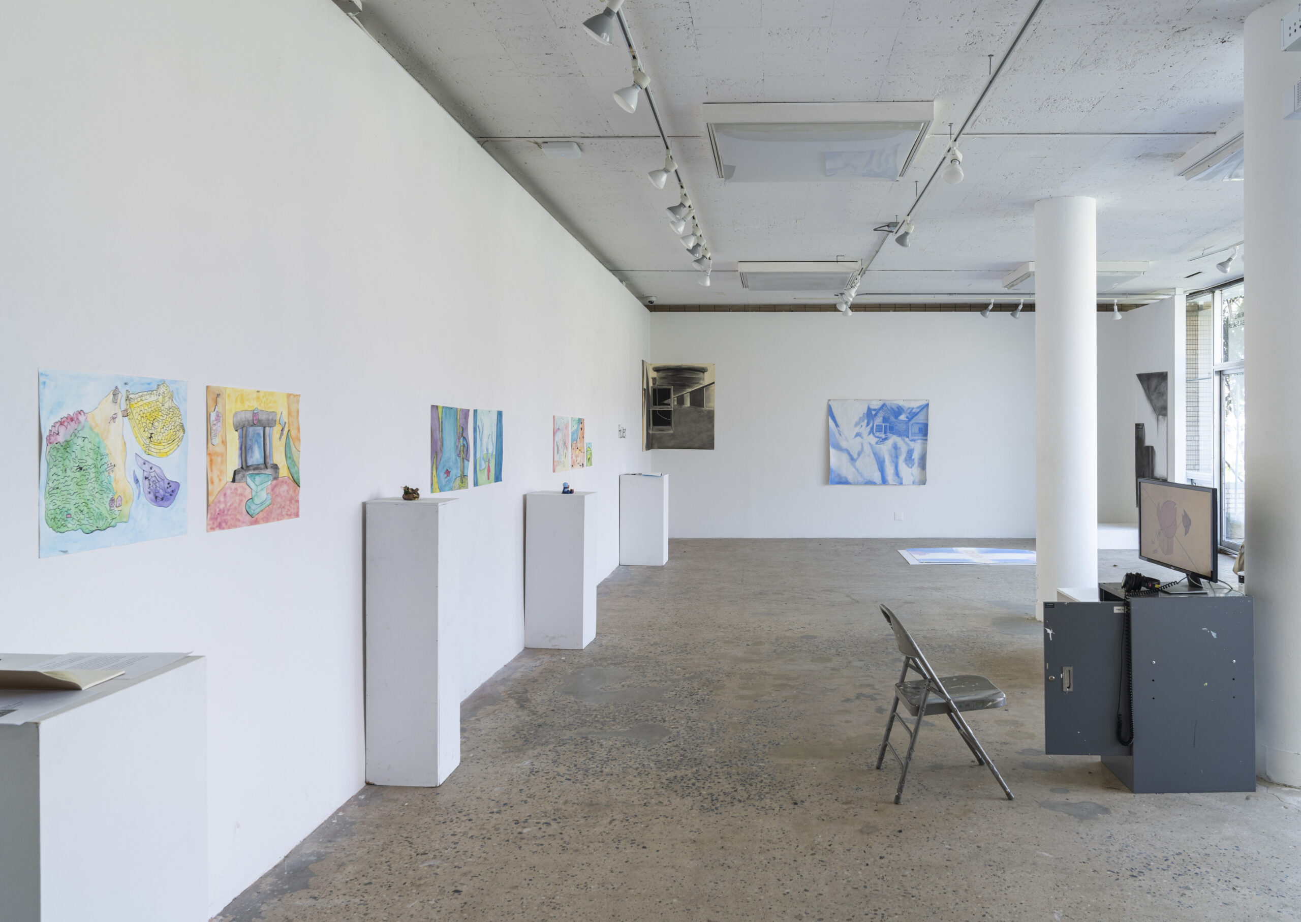 A brightly lit gallery featuring on the left wall: drawings unframed installed on the wall, alternating with pedestals atop which are miniature sculptures. On the corner is installed a black and grey drawing on the wall. The back wall hangs a blue and white drawing. In the center of the room is a folding chair and metal cabinet, which has a desktop computer on it.