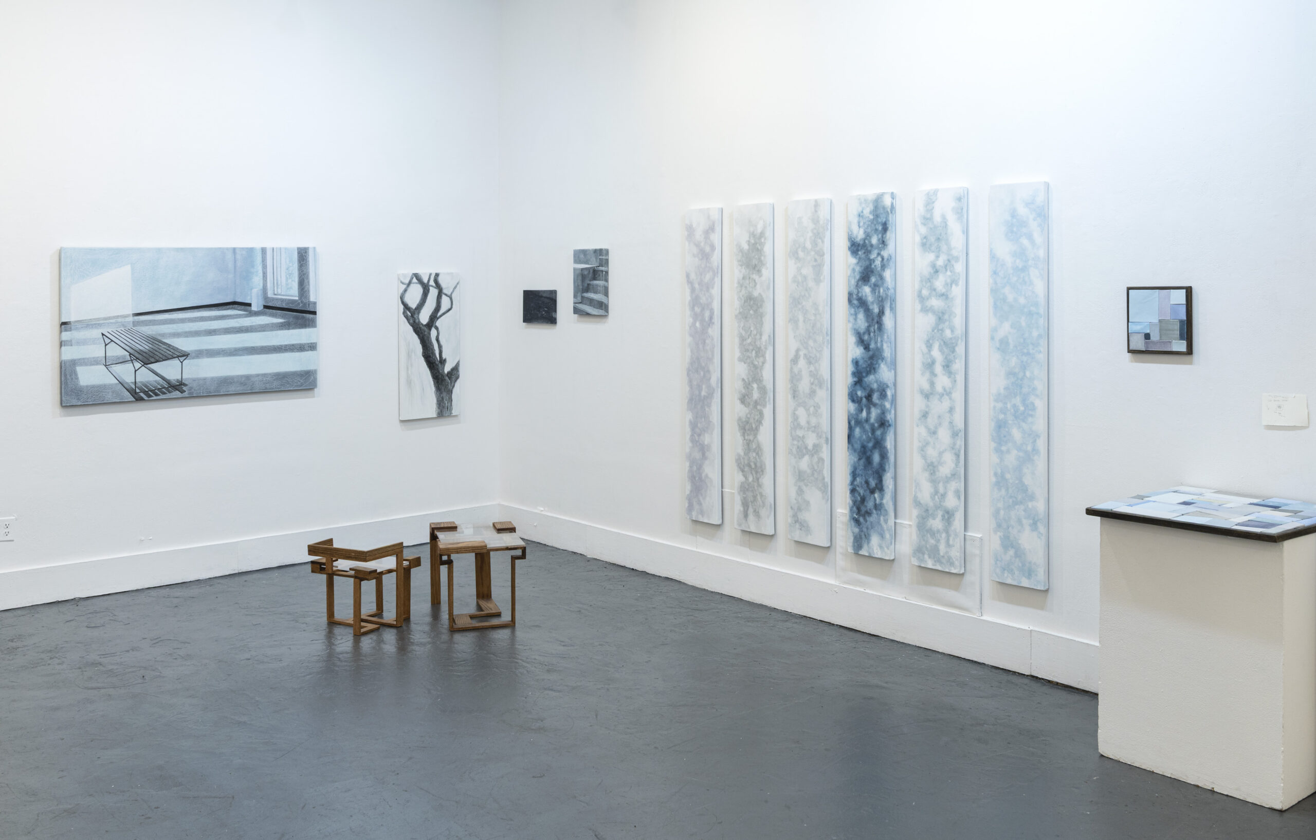 A brightly lit gallery with grey floors and white walls. On the walls hang blue and white paintings: two single paintings, one rectangular and the other amorphous; two small paintings in the corner; and a row of vertical hanging scrolls. Another painting rests face-up on a pedestal.