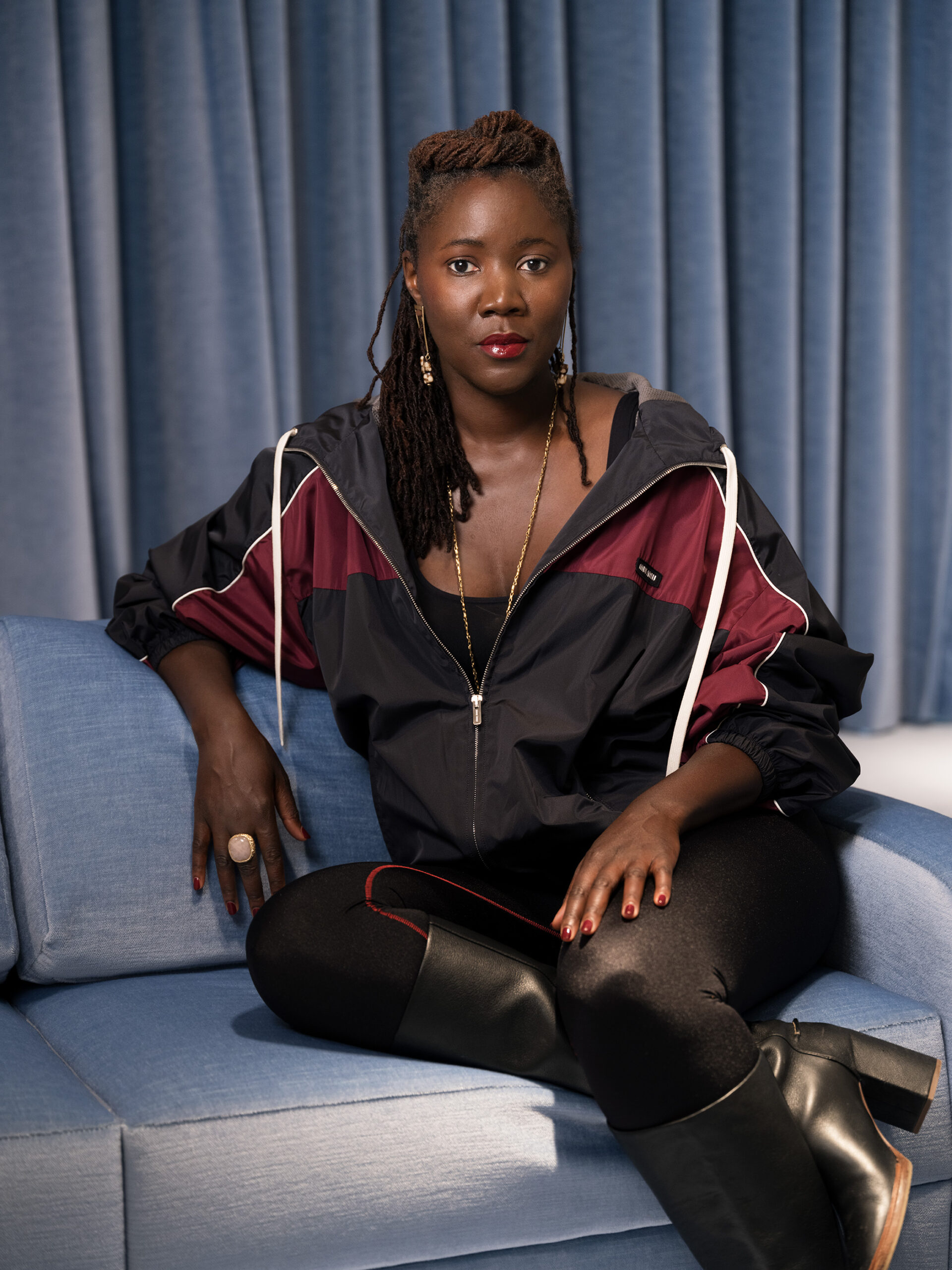 A portrait of a person seated on a light blue sofa in front of blue curtains. They wear a black and burgundy windbreaker, black leggings, and knee-high black boots. Their expression is calm and confident, with hands resting naturally on the sofa and knee, conveying a poised, introspective presence.