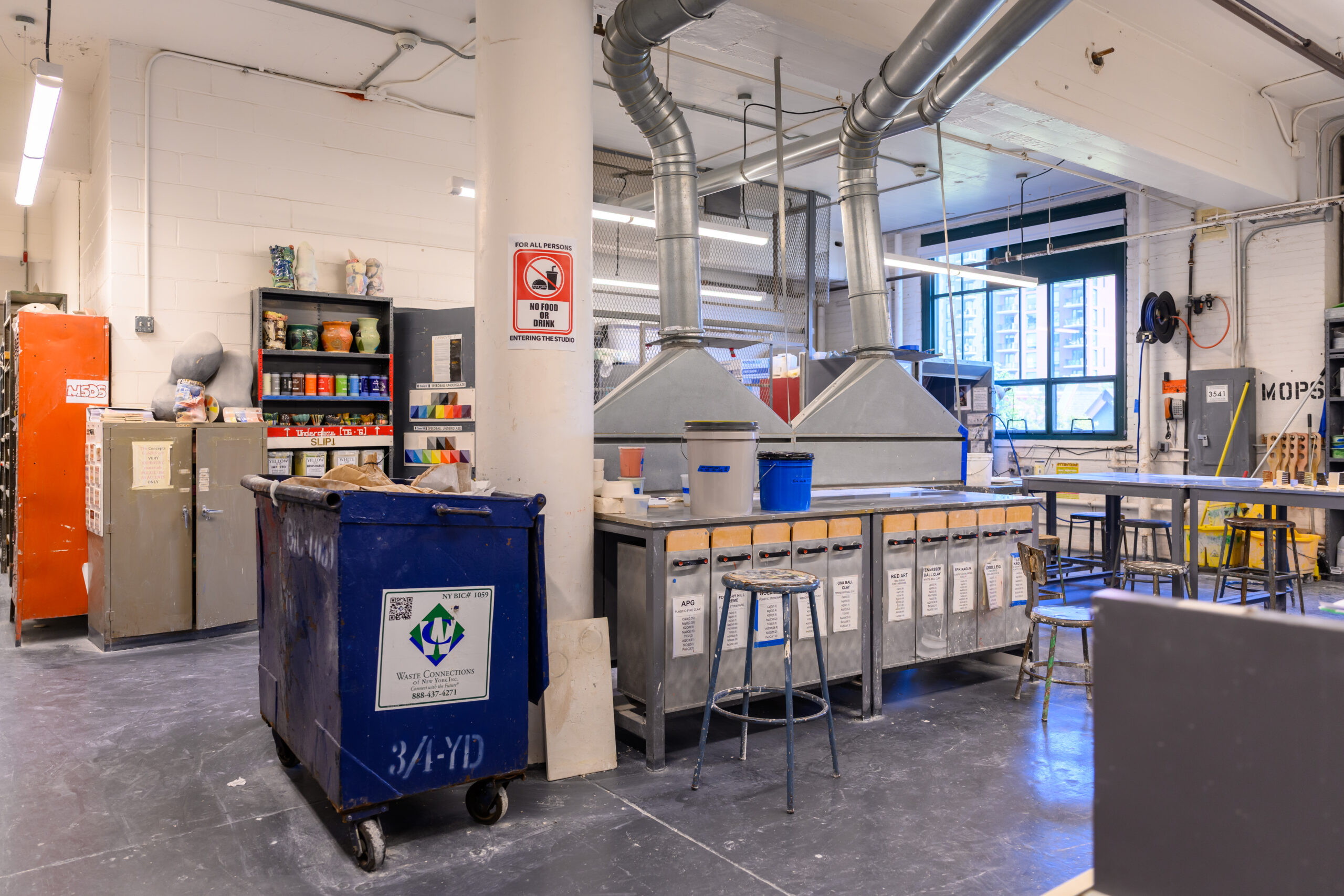 A photo of Fine Arts ceramic shops, featuring a shelf of varying slips, a trash bin, stools, tables, storage for different clays, and vent hoods, among other things.