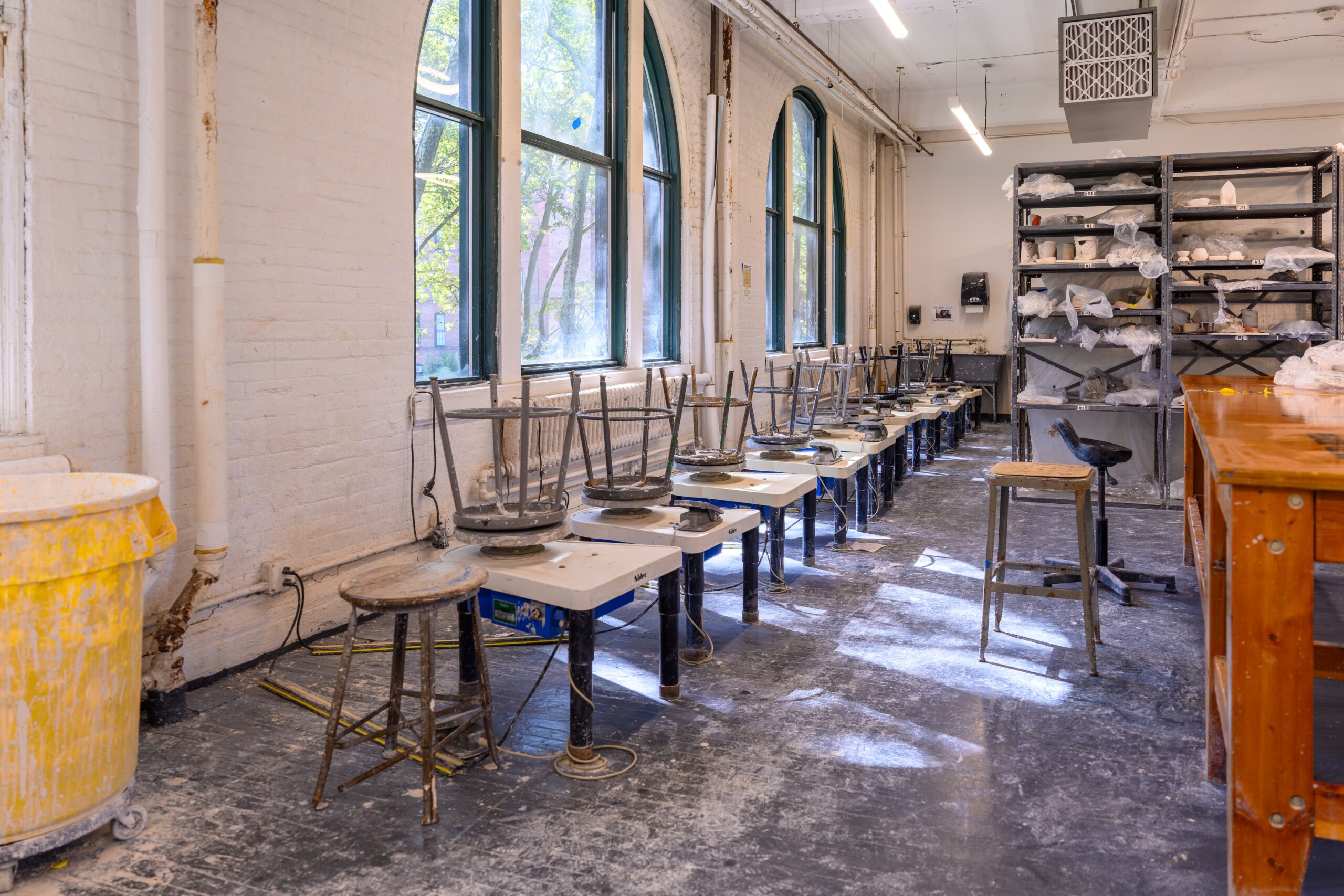 A row of throwing wheels with stools on top in the Fine Arts ceramics shop. In the background are shelves with ceramics pieces covered in plastic, drying in preparation to be fired.