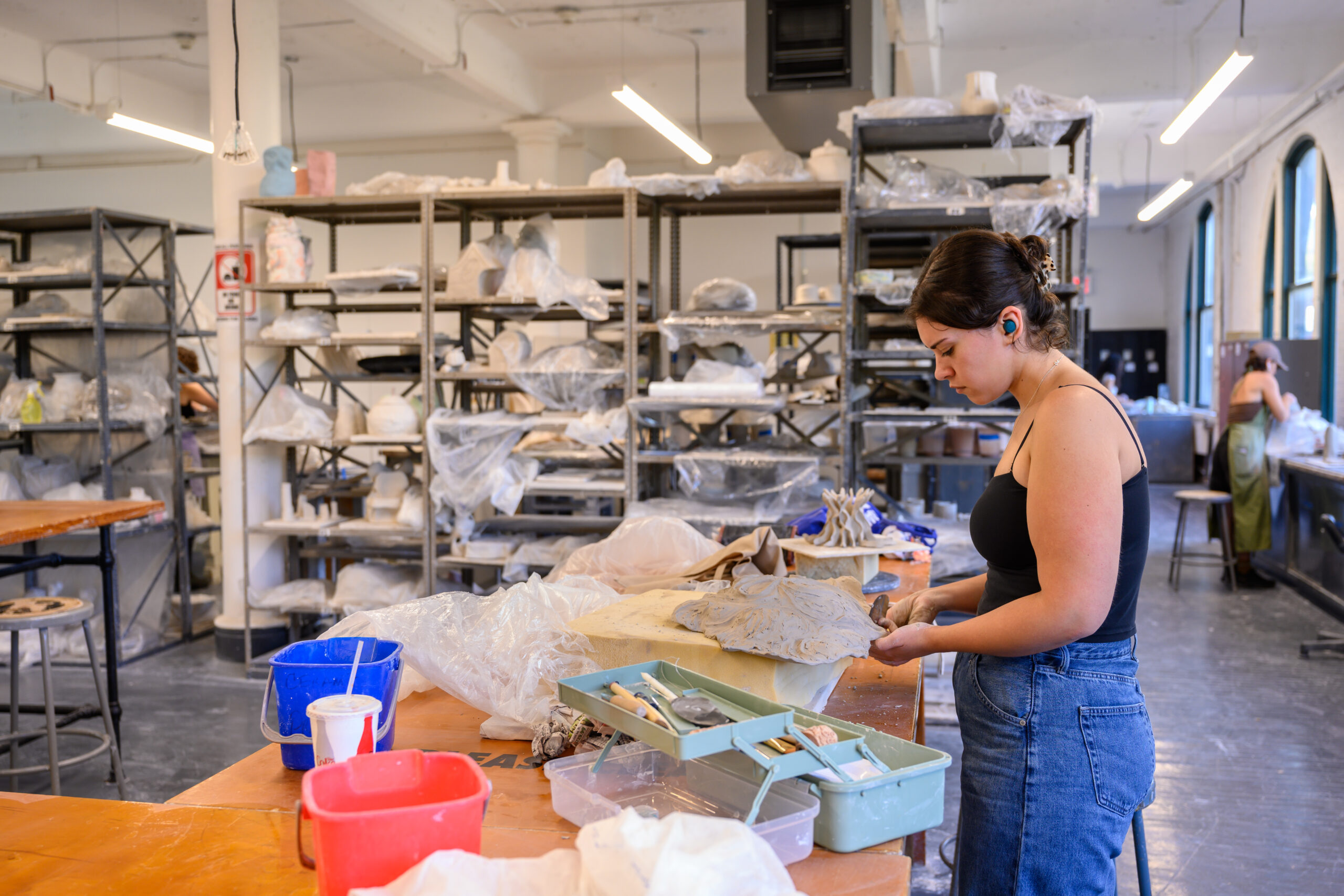 A student working on clay work at a table in the Fine Arts ceramic shop. Behind them are tall shelves with ceramic works covered in plastic, drying in preparation to be fired.