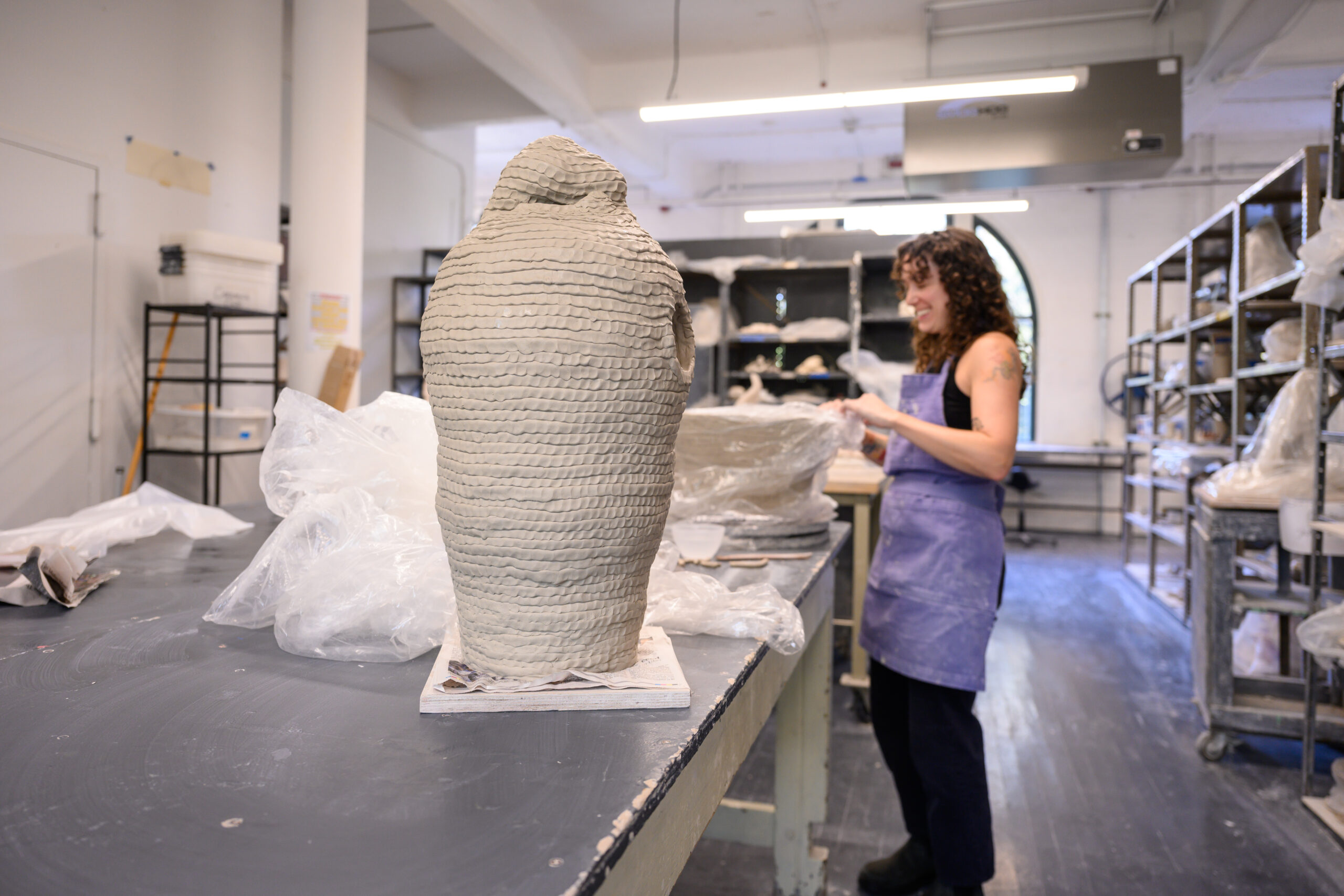 A student working on a tall coil-build vessel on a table in the Fine Arts ceramics shop