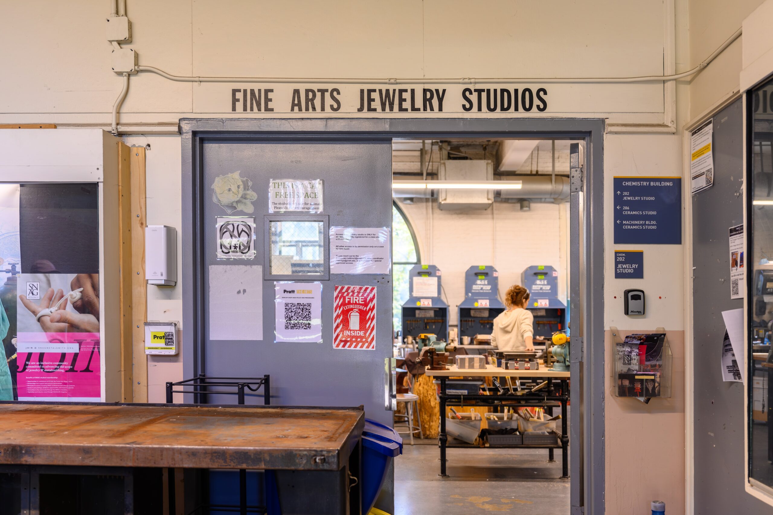 Entryway to Fine Arts Jewelry Studios. One door is shut, with postings on it for safety and policies. The other door is open, showing the inside of the Jewelry shop and a student at work.